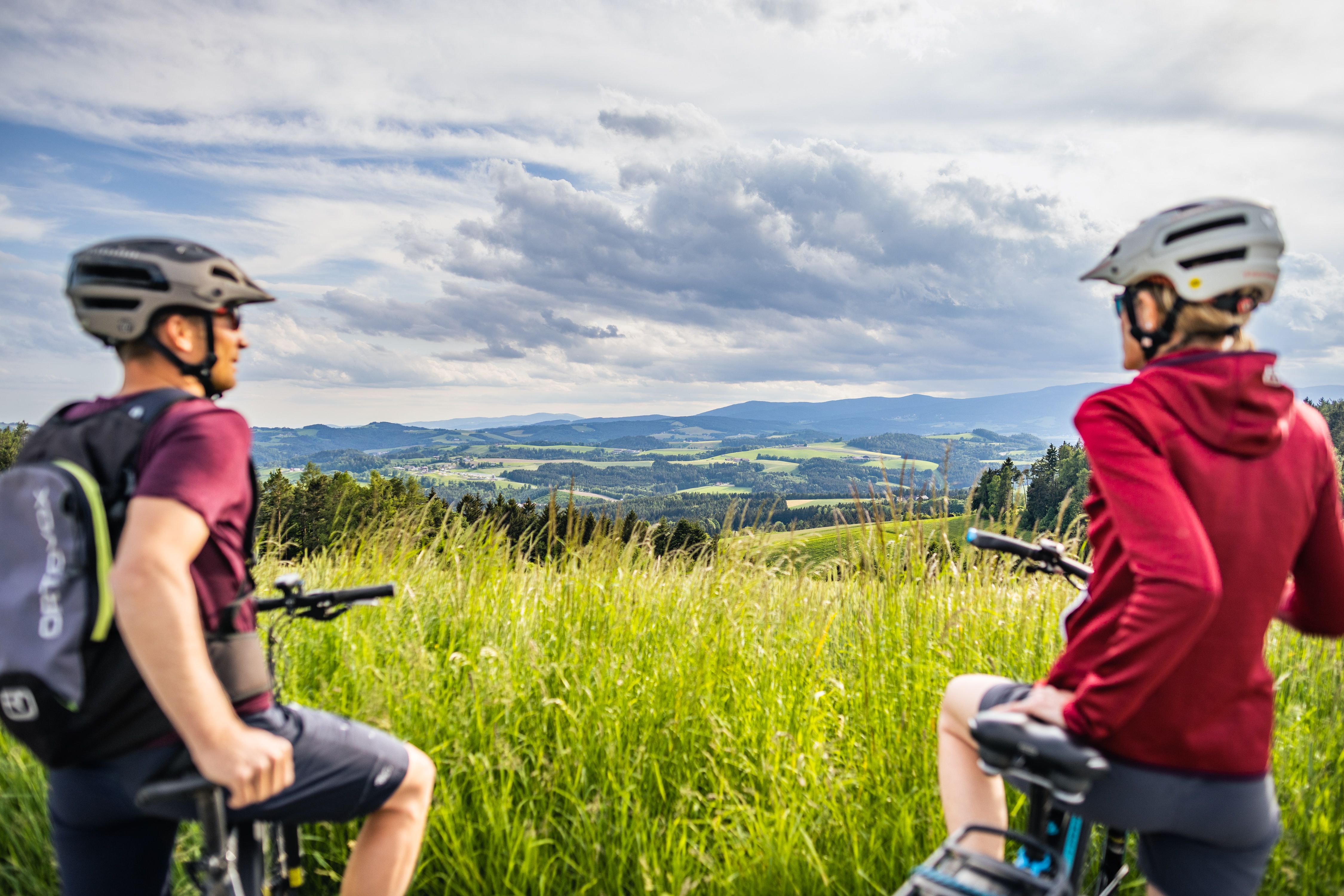Dva cyklisté s přilbami se dívají na kopcovitou krajinu luk a lesů pod zataženou oblohou.