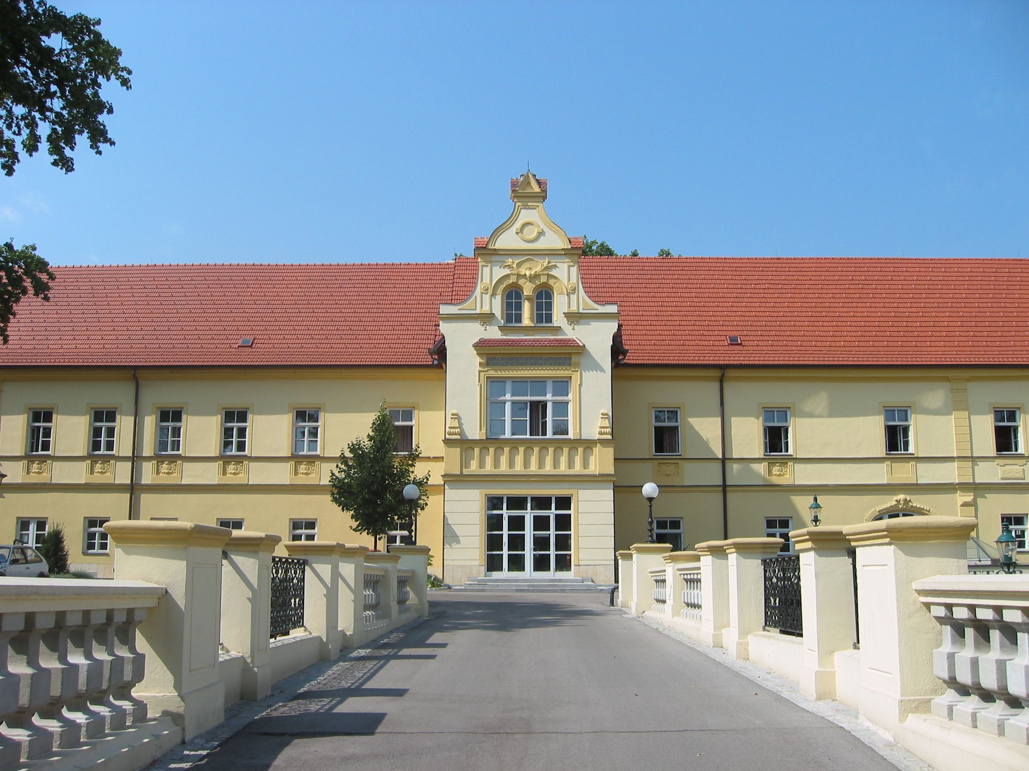 Žlutá budova s červenou střechou a ozdobnou fasádou, obklopená mostem a stromy.