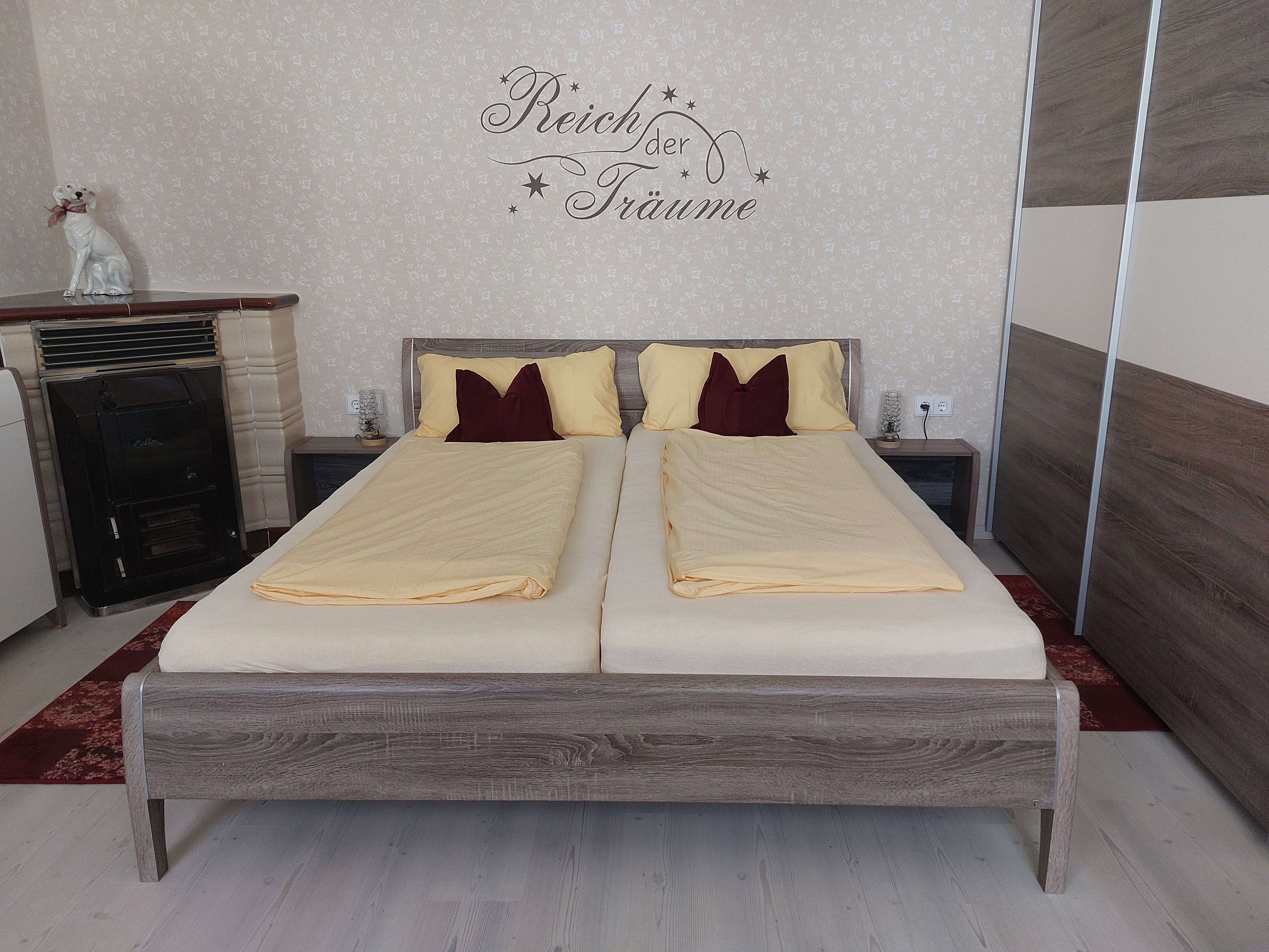 Ložnice s manželskou postelí, žlutým ložním prádlem a nápisem na zdi. K vidění jsou také kamna a socha psa.