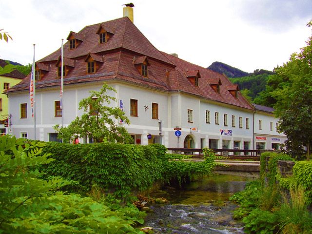 Historická budova s červenou střechou u potoka, obklopená zelení.