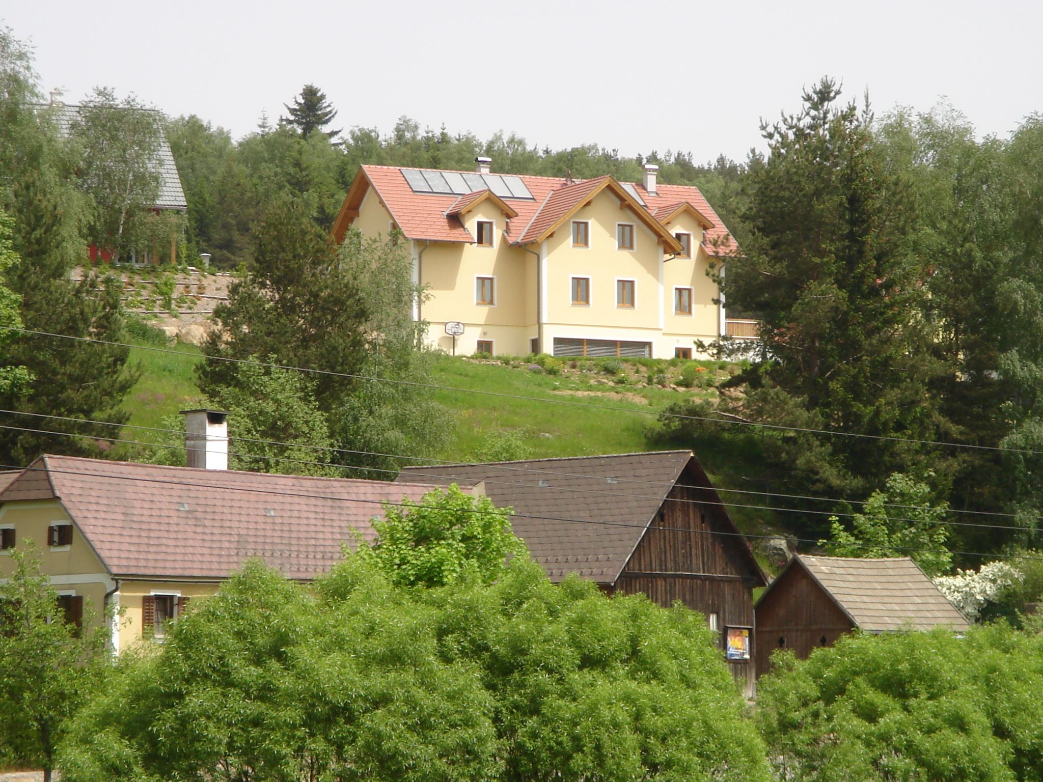 Na kopci stojí velký žlutý dům se solárními panely na střeše, obklopený stromy a menšími budovami v popředí.