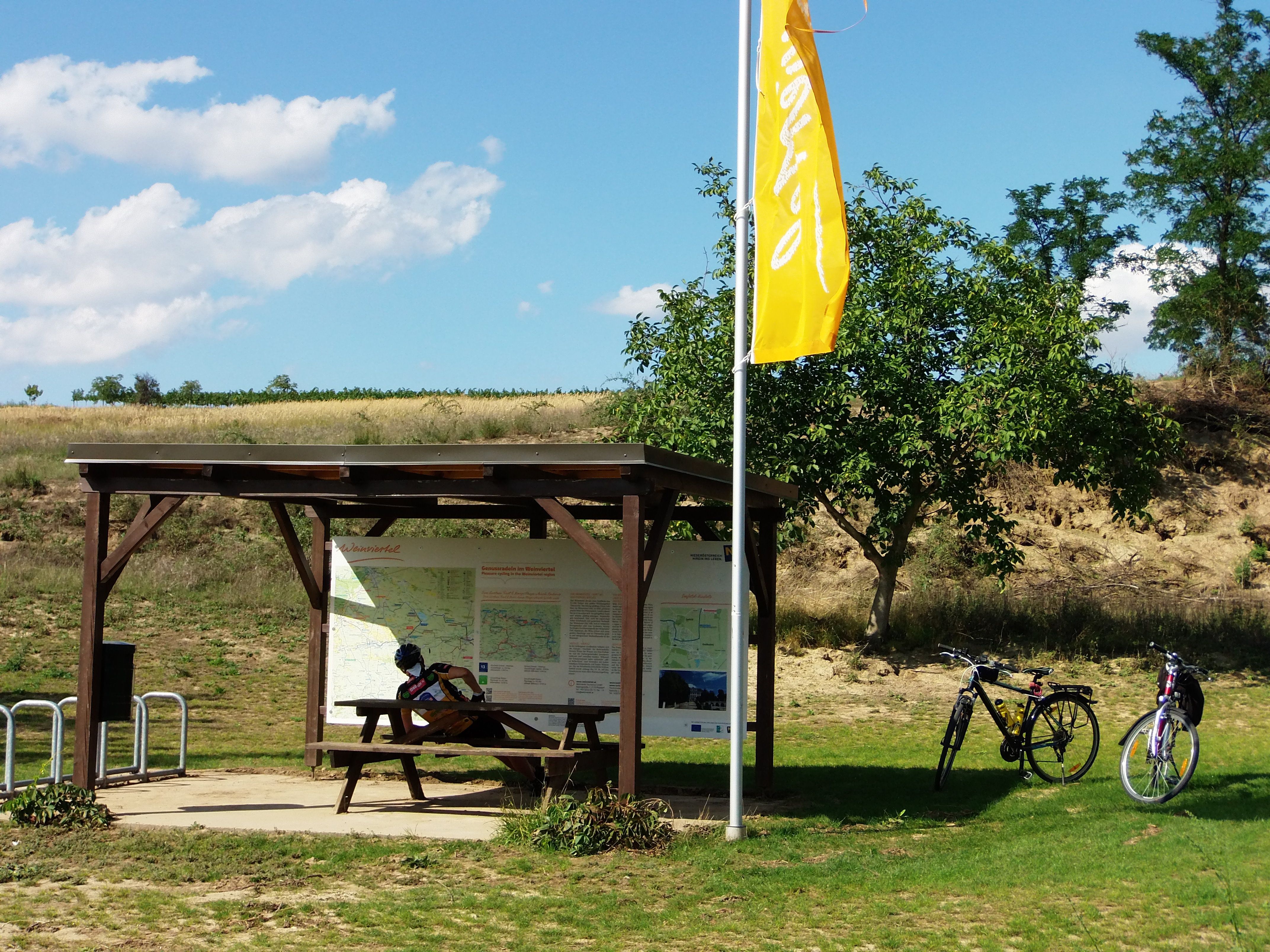 Odpočívadlo pro cyklisty s dřevěným stolem, dvěma koly a informační tabulí ve Weinviertelu.