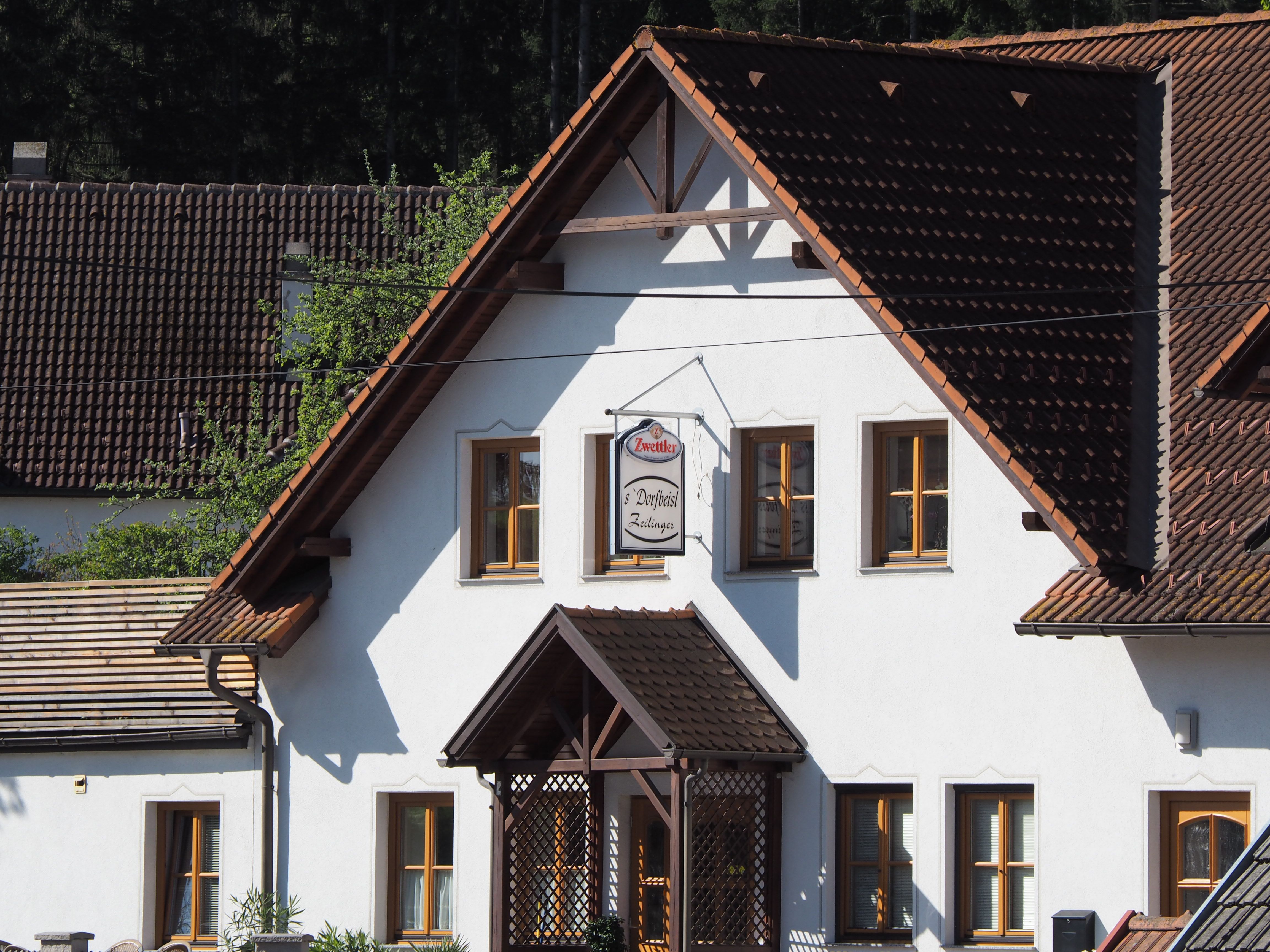Tradiční budova s bílými zdmi a hnědými střešními taškami. Na fasádě visí cedule s nápisem "Zwettler Dorfwirt Zeilinger".