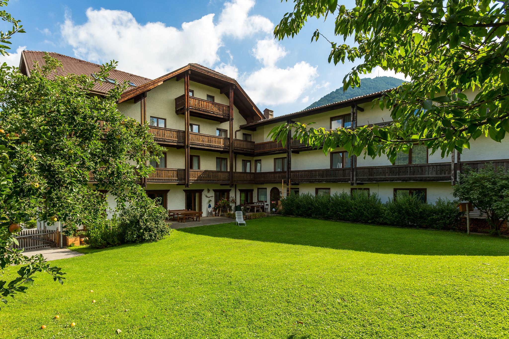Tradiční hotel s dřevěnými balkony a udržovanou zahradou v popředí, obklopený stromy a horami v pozadí.
