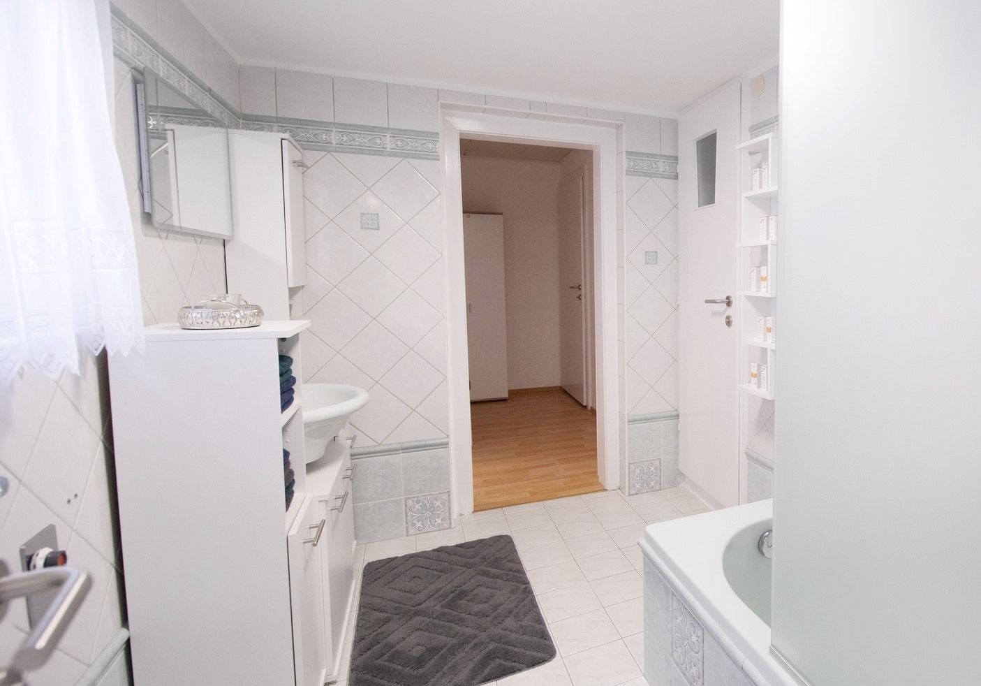 Moderní koupelna s bílými obklady, vanou, umyvadlem a skříňkou. Dveře vedou do předsíně s dřevěnou podlahou.