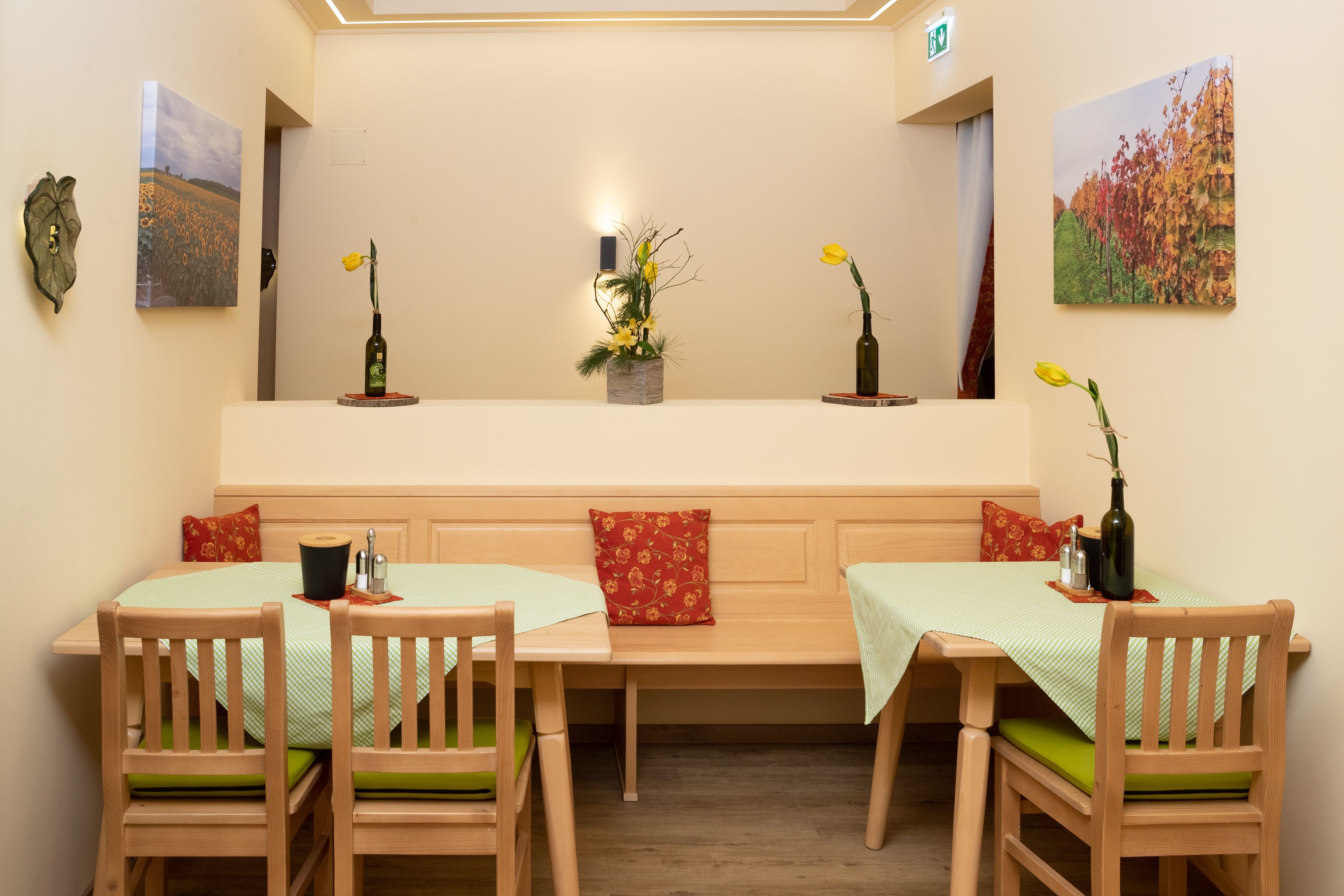 Útulná snídaňová místnost s dřevěným nábytkem, zelenými ubrusy a žlutými květinami ve vázách. Na stěnách visí obrazy krajin.