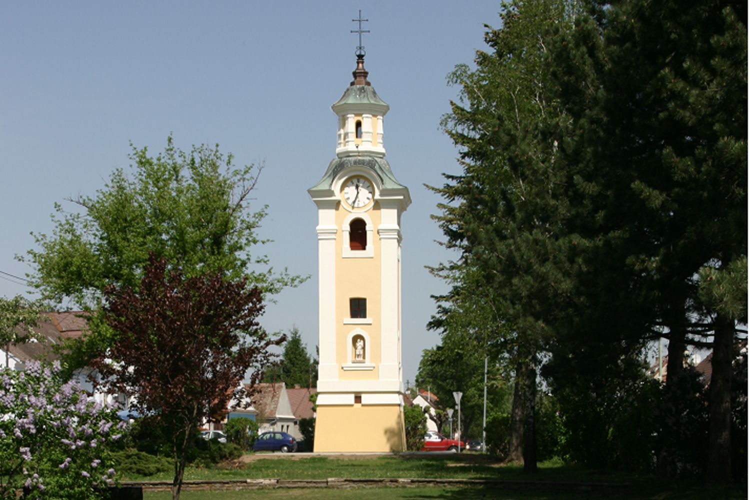 Žlutá kostelní věž s hodinami v Hohenau, obklopená stromy.