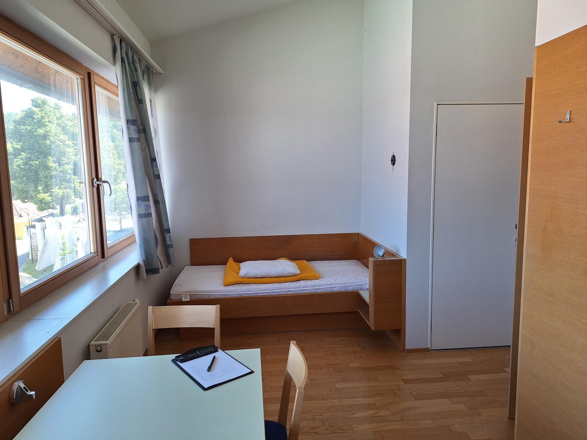 Jednoduše zařízený jednolůžkový pokoj s postelí, stolem a židlí, světlým dřevěným nábytkem a velkým oknem s výhledem ven.
