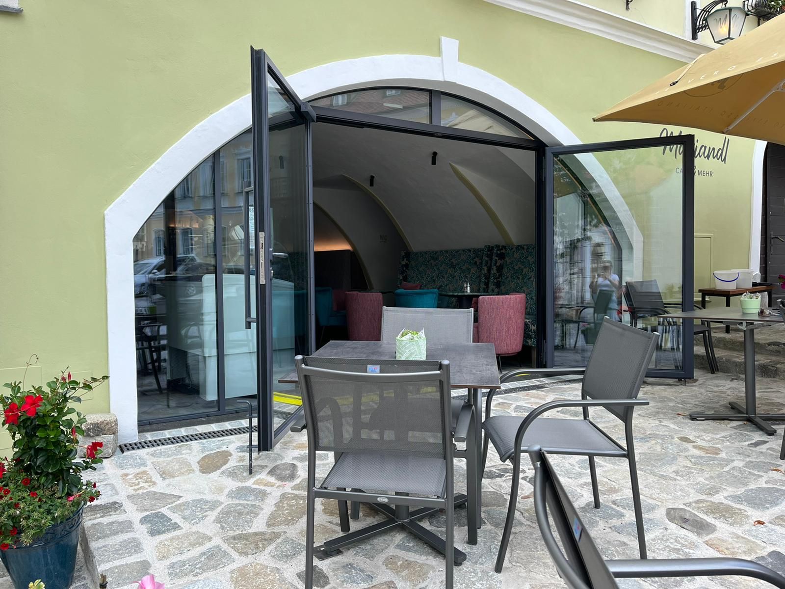 Pohled zvenčí na kavárnu s otevřenými prosklenými dveřmi a venkovním posezením.