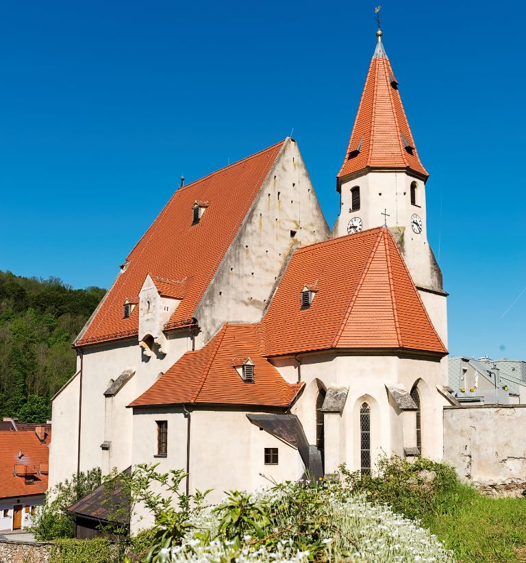 Opevněný kostel v Edlitz s mnoha bočními částmi, červenými taškovými střechami, špičatou věží a modrou oblohou.
