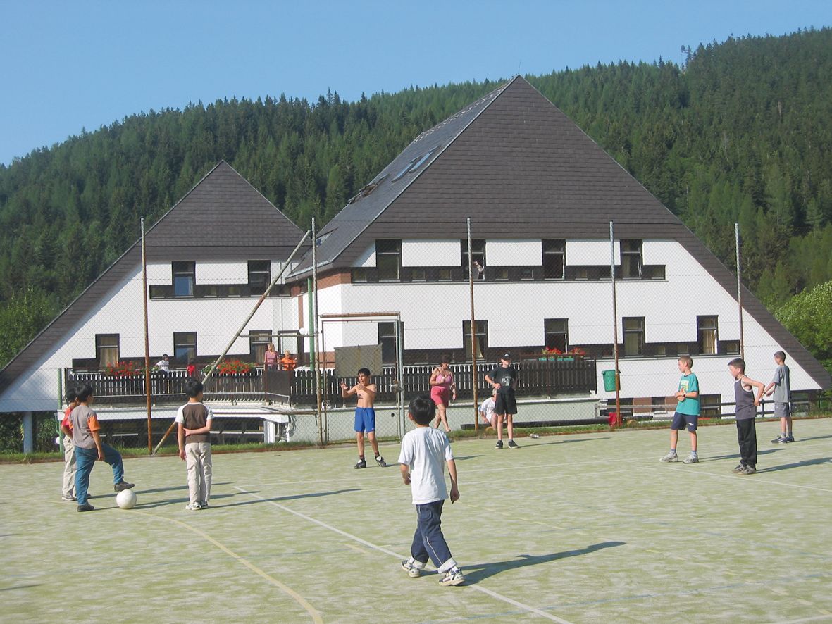 Děti hrají fotbal na hřišti před hotelem v zalesněné oblasti.