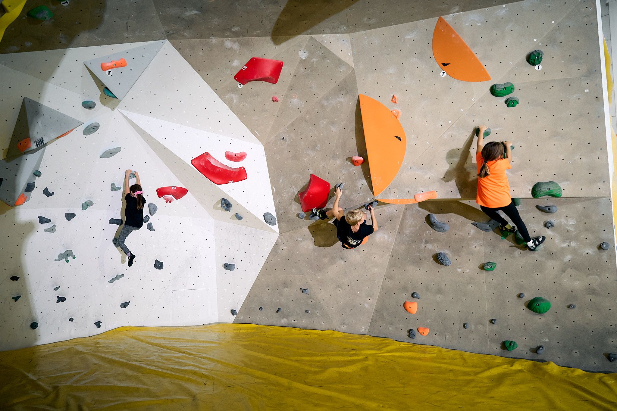 Tři děti lezou na kryté boulderingové stěně s barevnými chyty.
