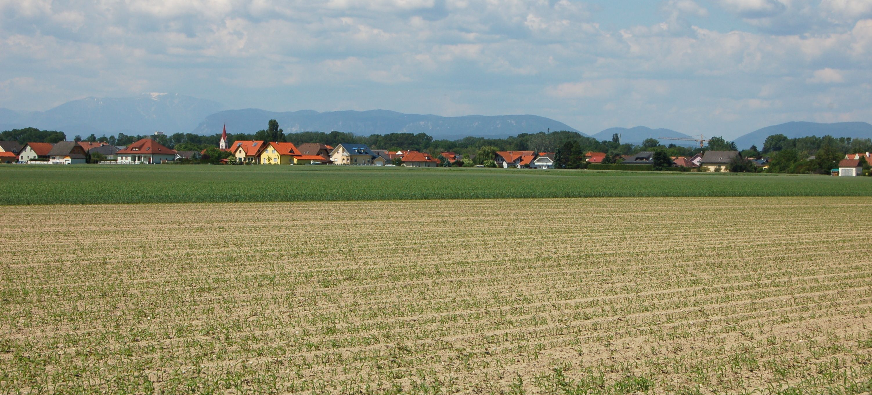 Krajina s poli a vesnicí v pozadí, hory na obzoru.