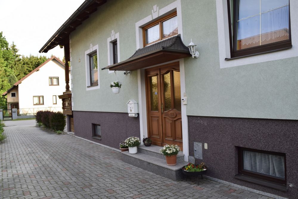 Vchod do domu se zelenou fasádou a dřevěnými dveřmi, obklopený rostlinami v květináčích.