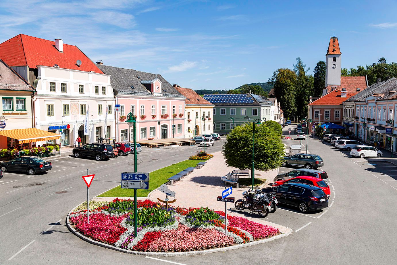 Hlavní náměstí Aspang Markt s barevnými květinovými záhony, zaparkovanými auty a historickými budovami.