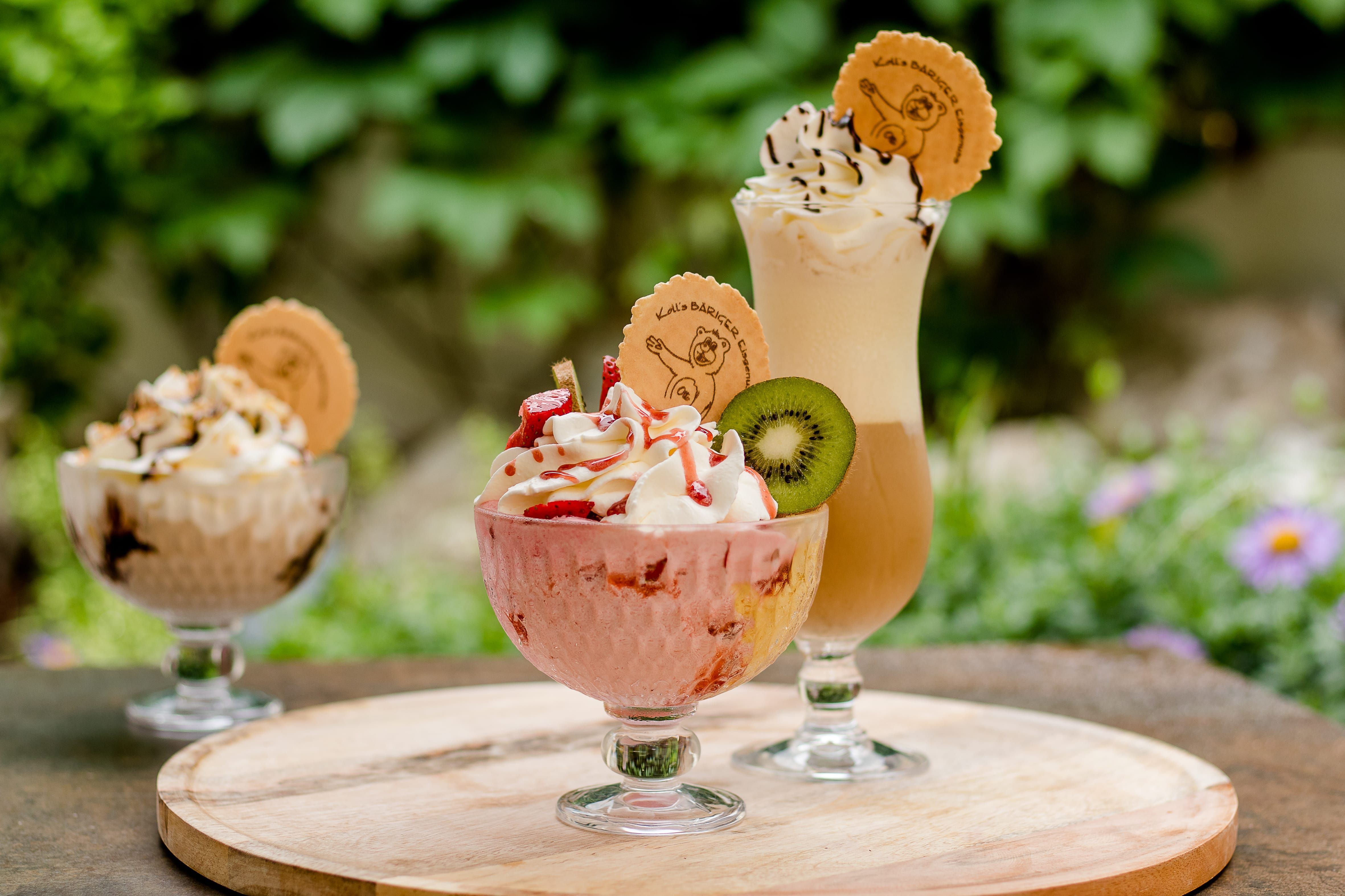 Tři poháry se smetanou a zdobením na dřevěném stole venku.