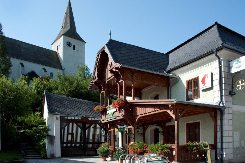 Tradiční budova s dřevěným balkonem a květinovou výzdobou, vedle kostela s věží.