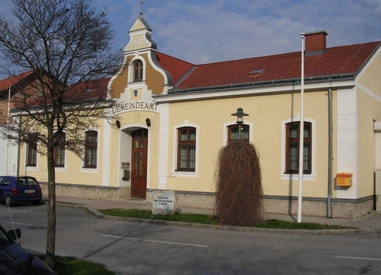 Žlutá budova s nápisem "Gemeindeamt", červenou střechou, stromem a lucernou před ní.
