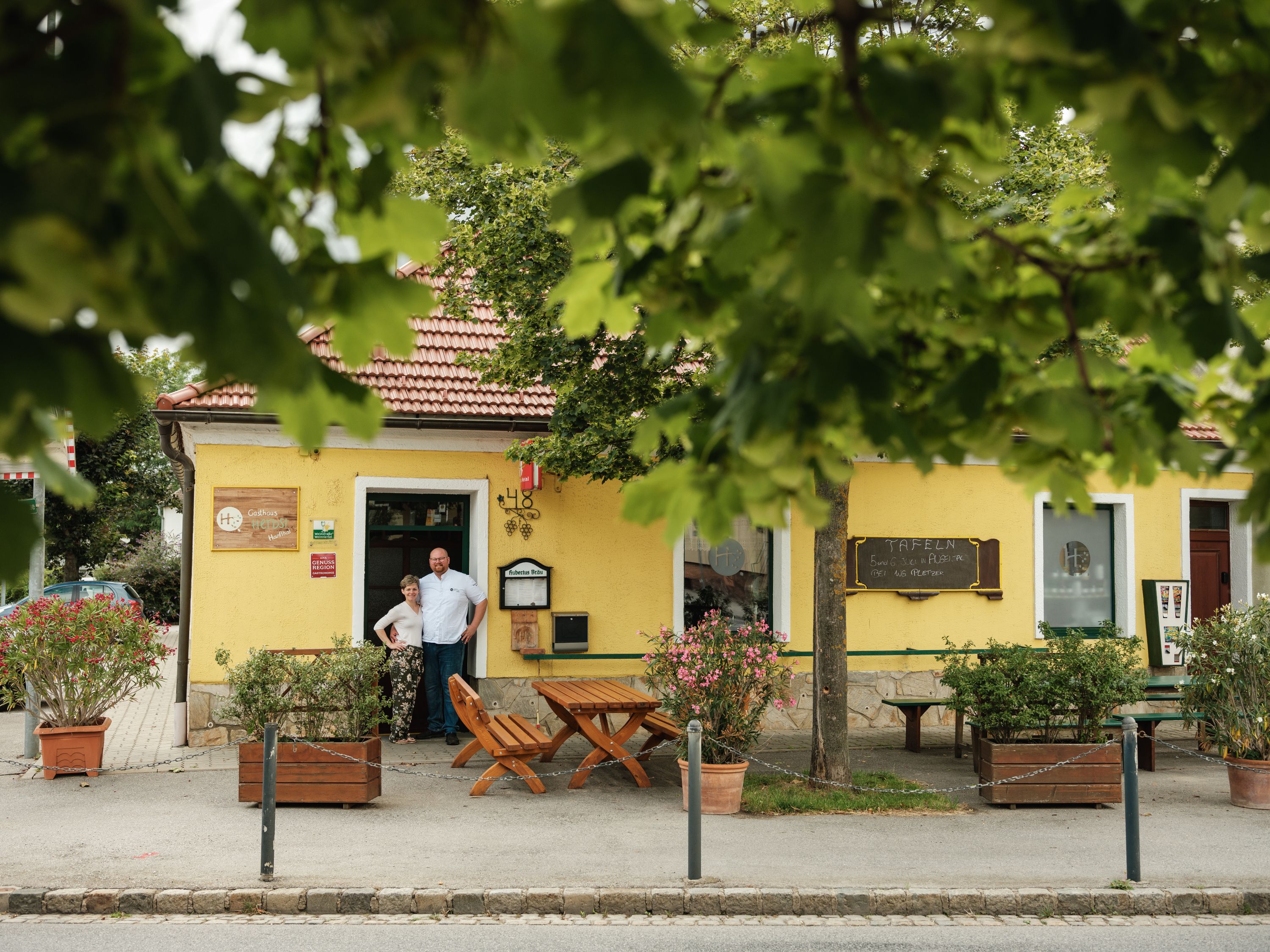 Žlutý hostinec s červenou střechou, obklopený stromy. Před vchodem stojí dva lidé.