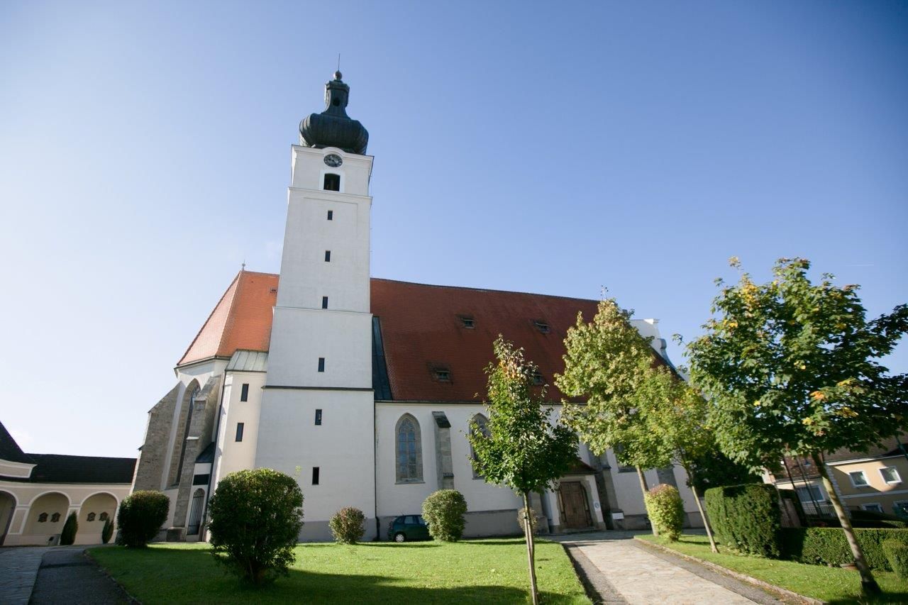 Mankův poutní kostel s věží a červenou střechou, obklopený stromy a trávníkem.