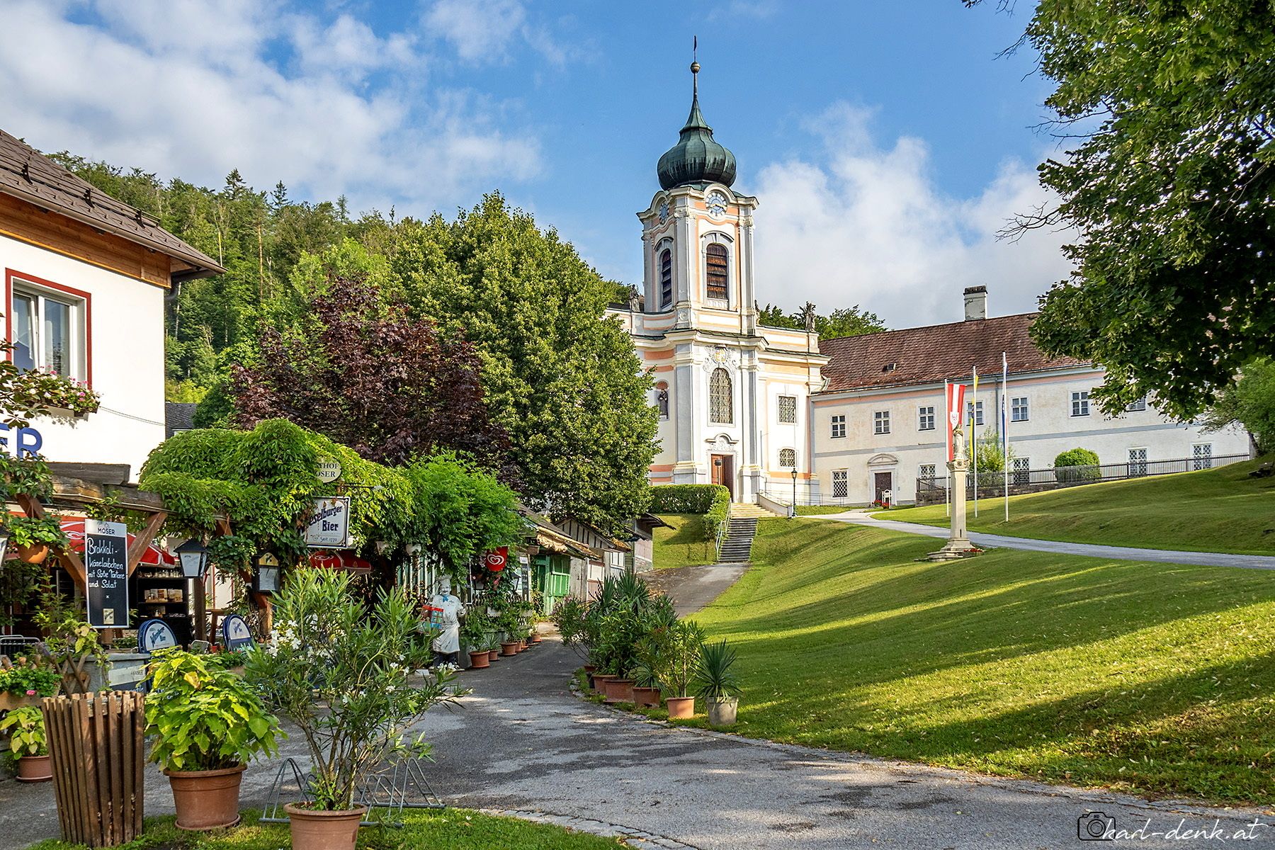 Malebná vesnice s barokním kostelem a hostincem v popředí, obklopená zelenými stromy a modrou oblohou.