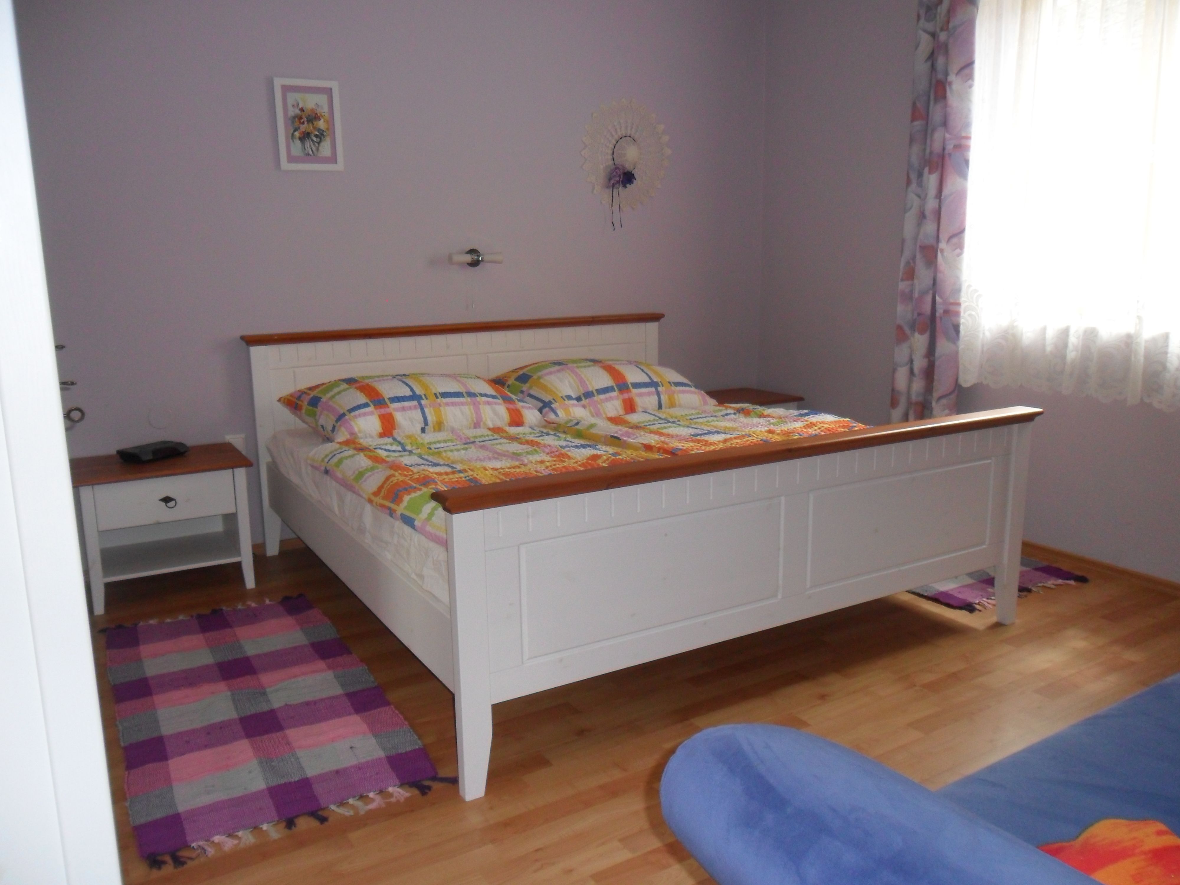 Ložnice s bílou postelí, barevnými peřinami, nočním stolkem a oknem se závěsy.