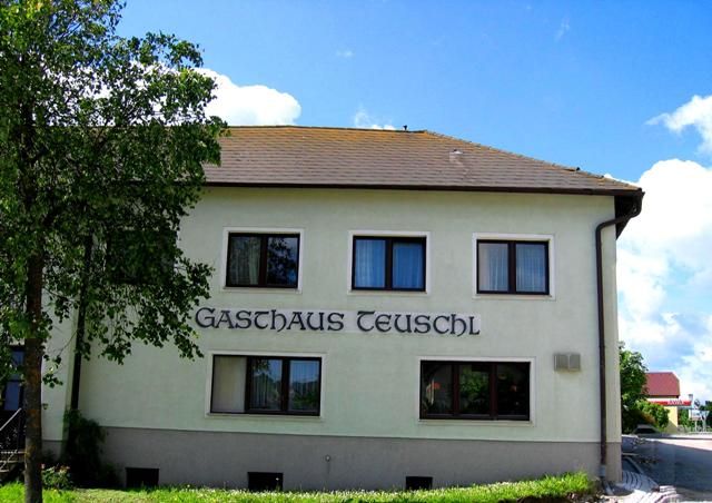 Pohled zvenčí na hostinec Teuschl se stromy a modrou oblohou.