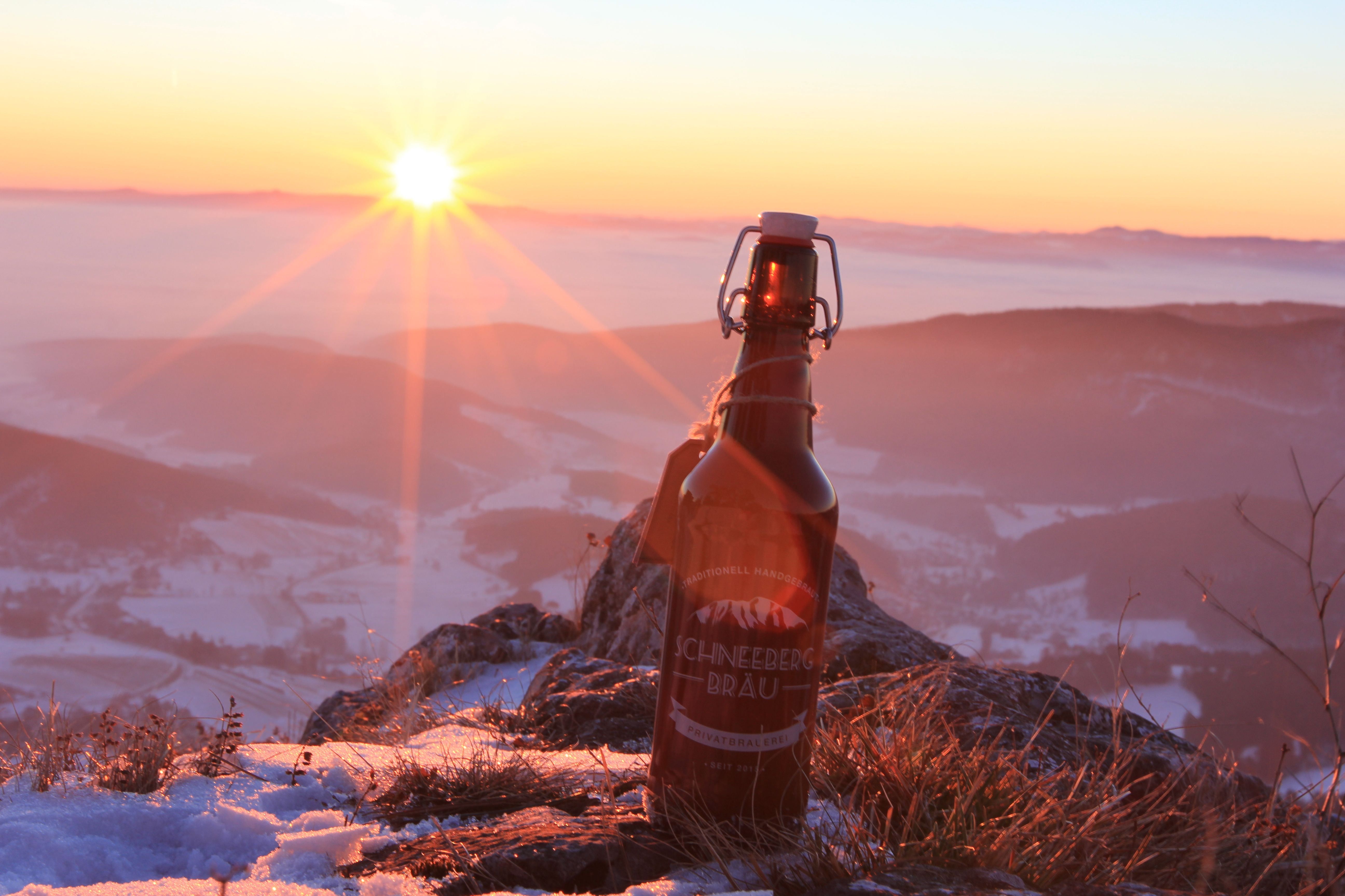 Láhev piva Schneeberg Bräu stojí na zasněžené hoře při západu slunce.