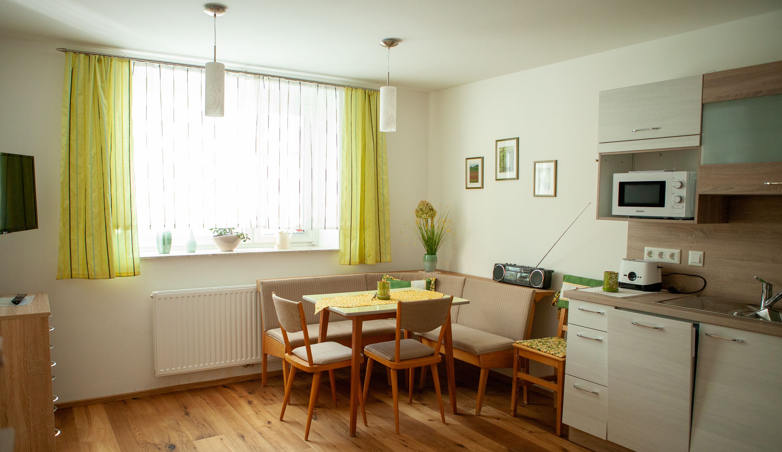 Útulná kuchyně s jídelním stolem, rohovou lavicí, kuchyňskou linkou a zelenými závěsy.
