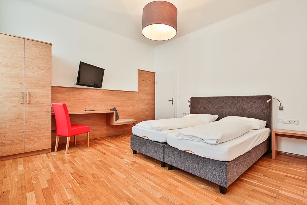 Hotelový pokoj s manželskou postelí, červenou židlí, psacím stolem a televizí.