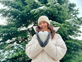 Žena v b&iacute;l&eacute;m kab&aacute;tě drž&iacute; před zasněžen&yacute;m stromem srdce ze sněhu.