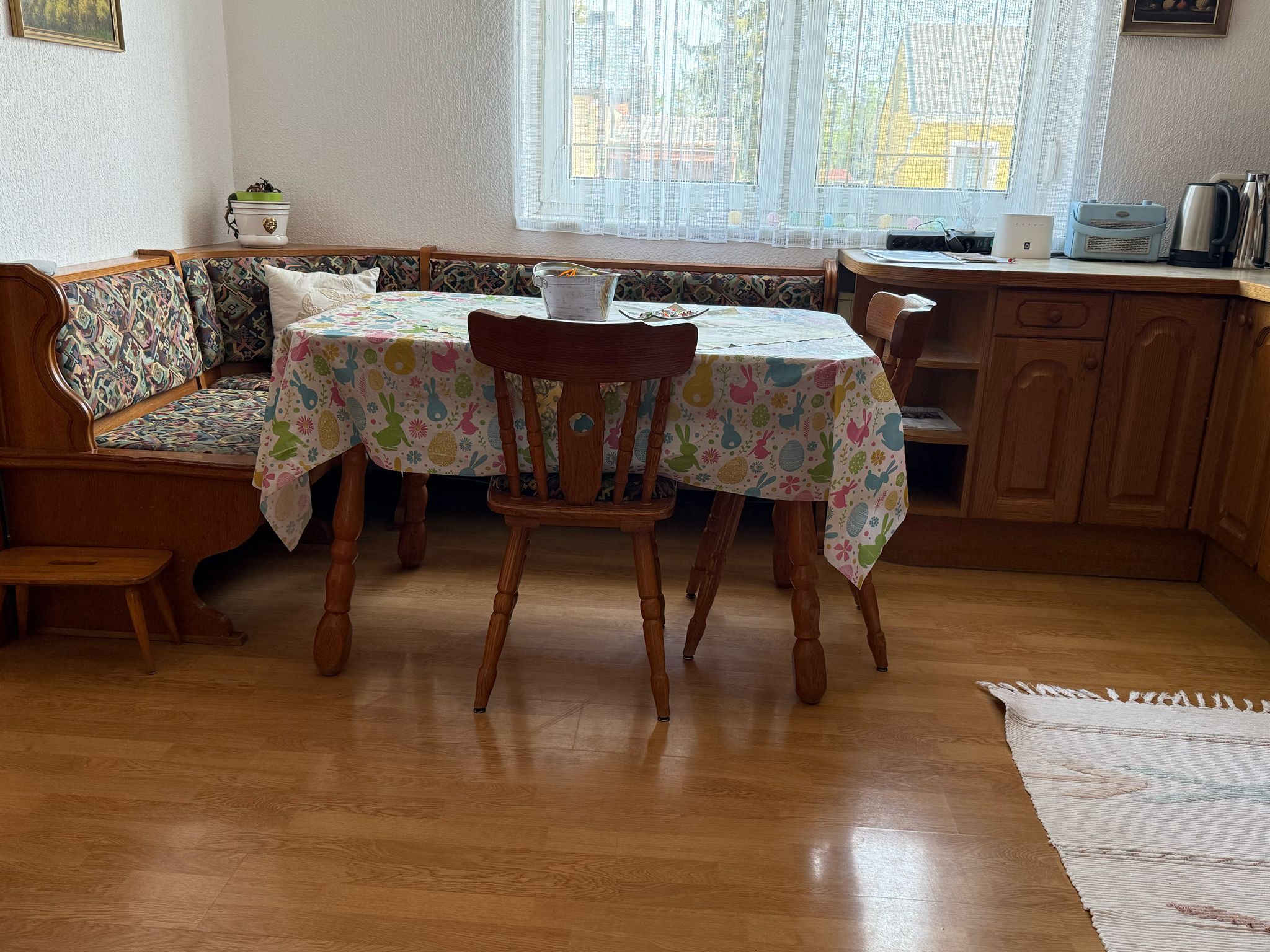Jídelní kout s dřevěným stolem, židlemi a rohovou lavicí, barevný ubrus, okno v pozadí.