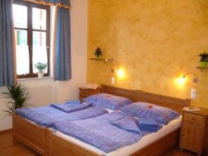 Útulný dvoulůžkový pokoj s dřevěným nábytkem, modrým ložním prádlem a žlutou stěnou. Jsou vidět rostliny a okna s modrými závěsy.
