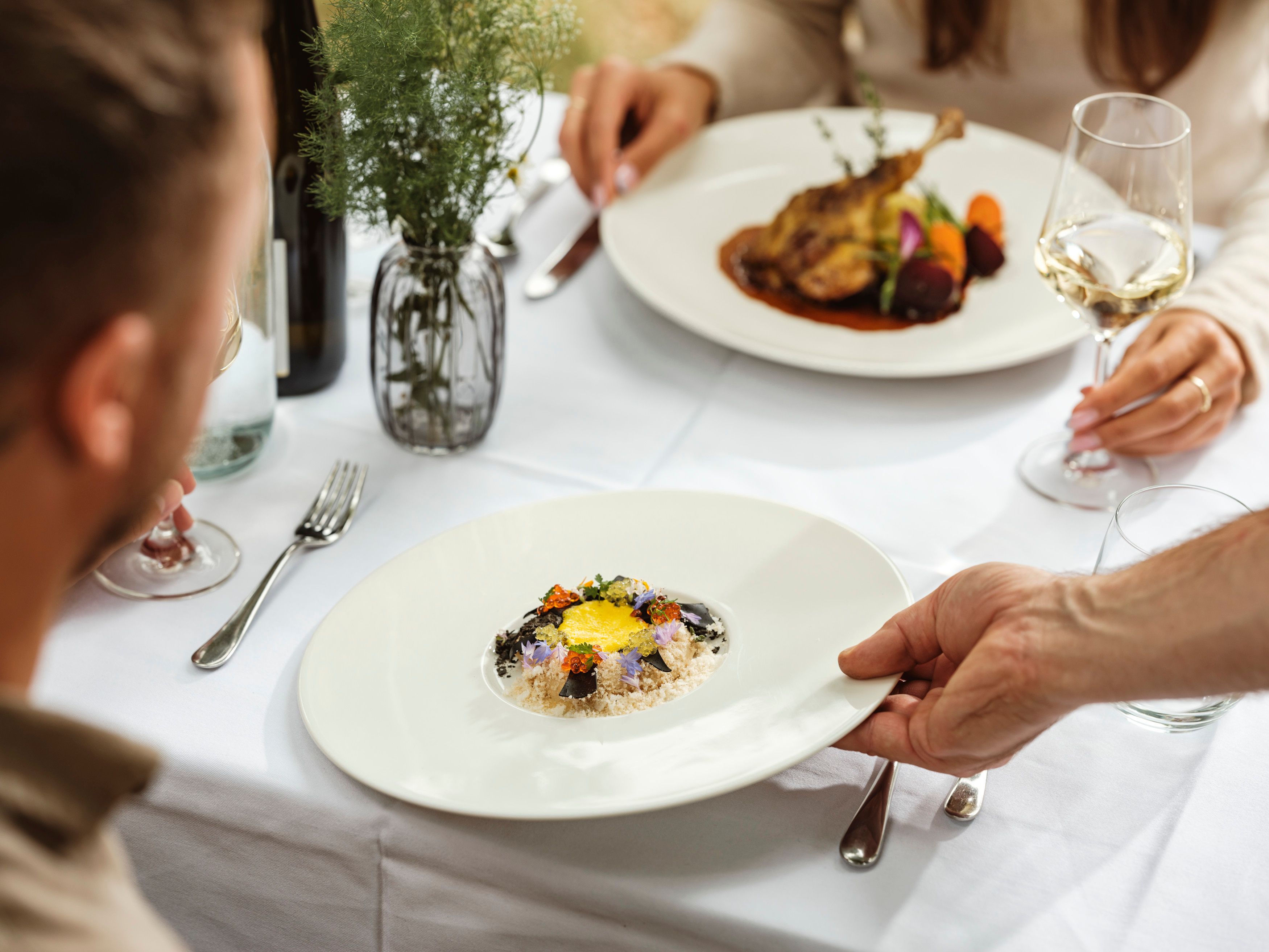 Elegantní jídlo v restauraci, kde u jednoho stolu sedí dvě osoby. Podává se talíř s umně připraveným pokrmem.