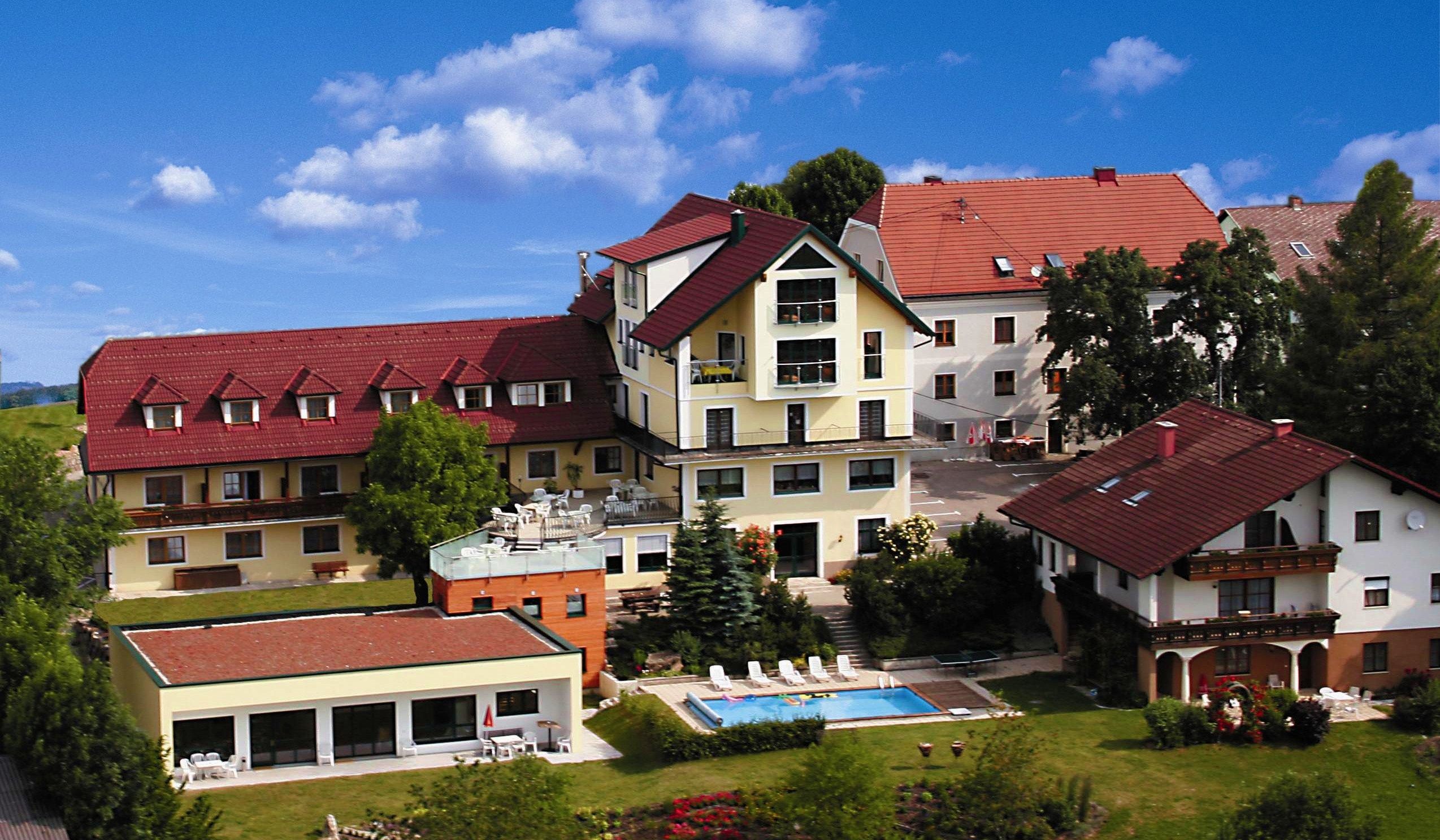 Hotelový komplex s červenými střechami, bazénem a zahradou, obklopený stromy a kvetoucími rostlinami pod modrou oblohou.