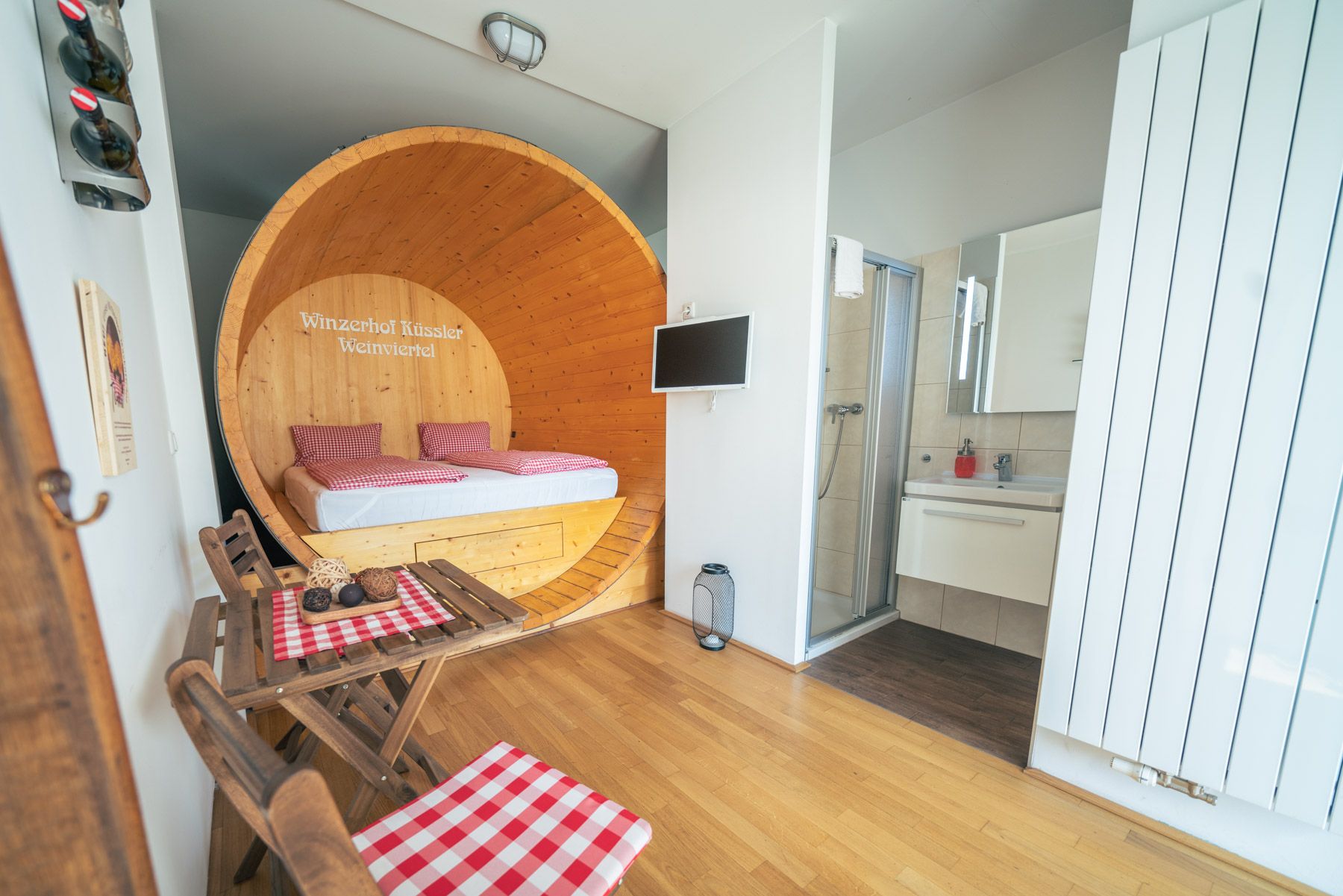 Pohled do interiéru apartmá s postelí ve velkém sudu na víno, malým stolkem se židlemi a koupelnou.