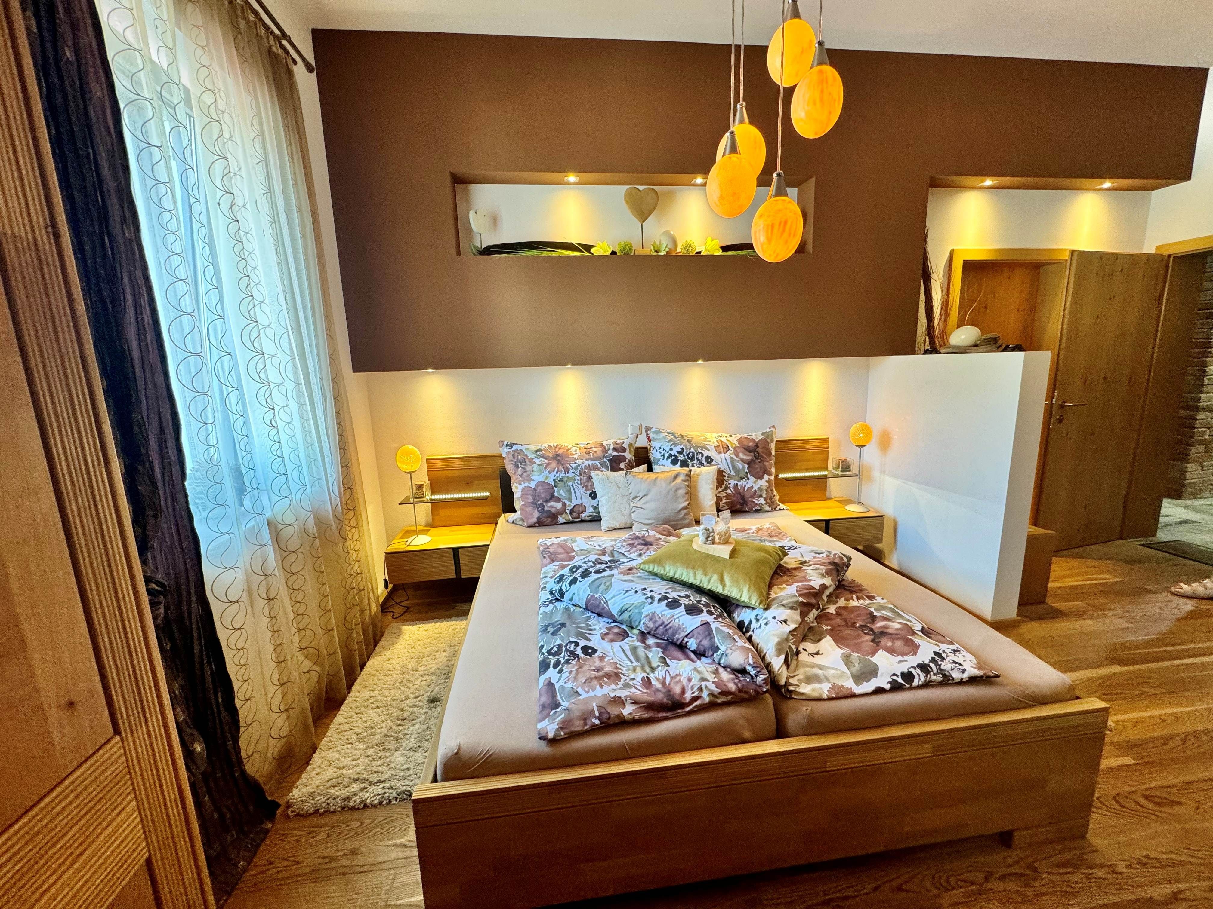 Útulná ložnice s manželskou postelí, ozdobnými polštáři a teplým světlem, které prostorem rozlévá světlo z lamp ve tvaru pštrosích vajec.