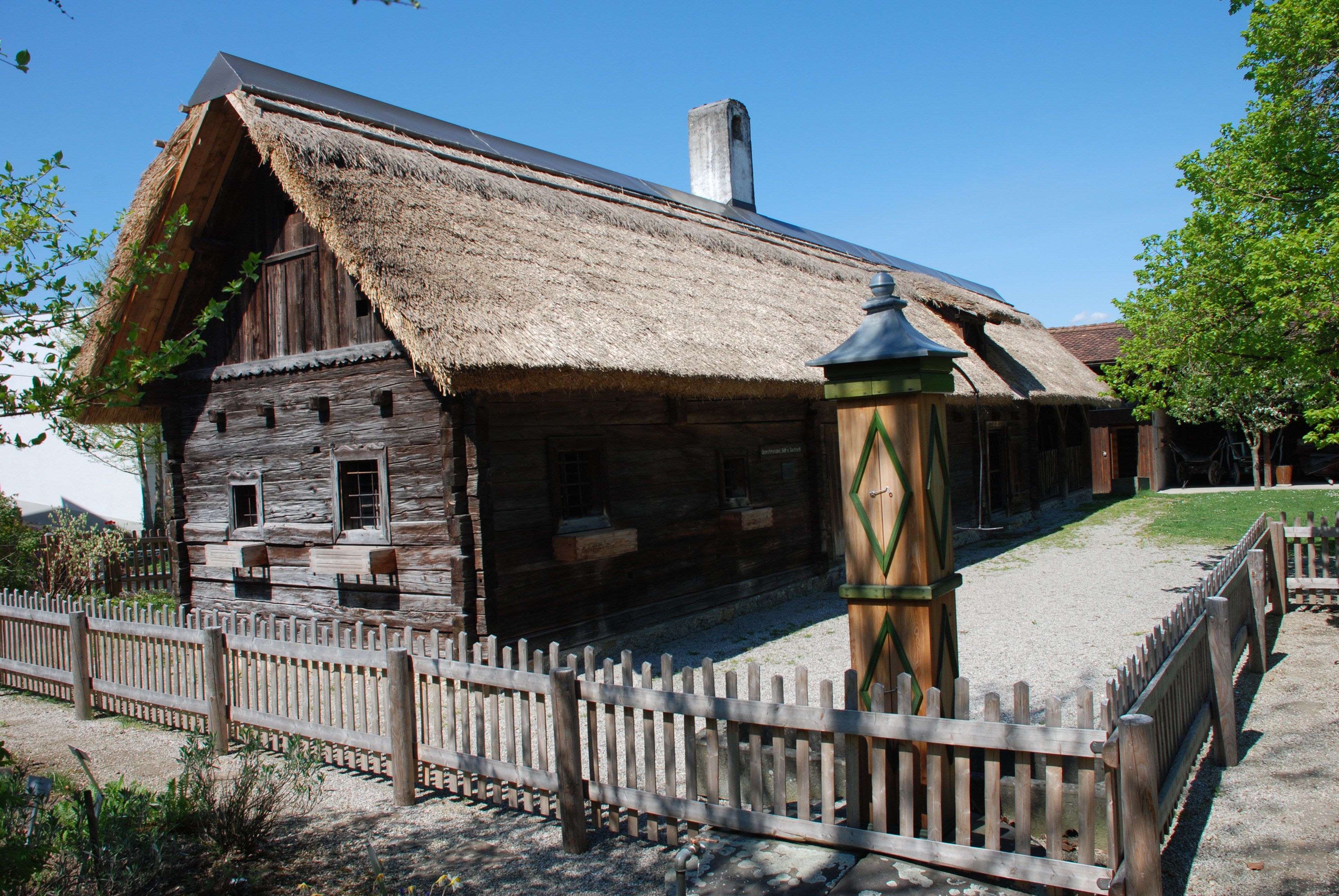 Tradiční dřevěný dům s doškovou střechou a dřevěným plotem ve venkovském prostředí.