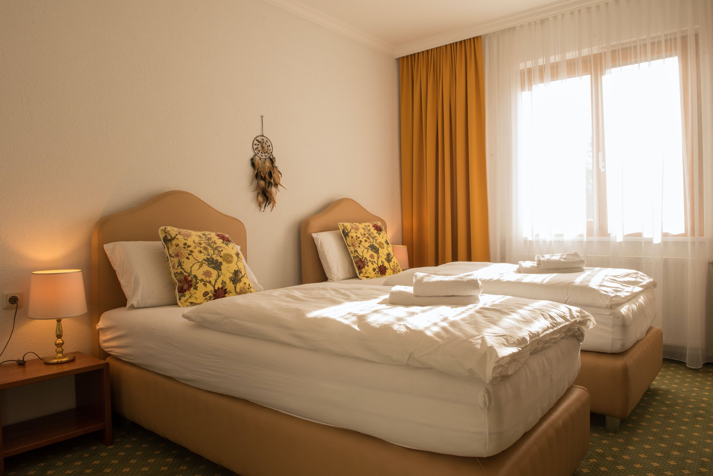 Útulný dvoulůžkový pokoj se dvěma samostatnými postelemi, žlutými závěsy a lapačem snů na zdi.