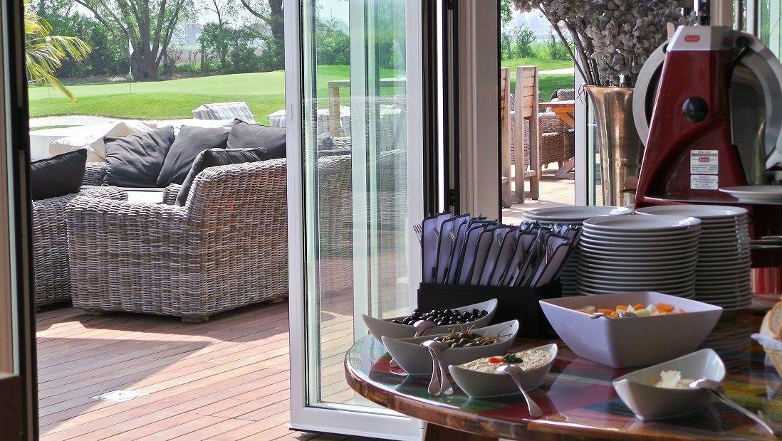 Stůl s nádobím a občerstvením, vedle terasa s ratanovým nábytkem a výhledem na zelený trávník.