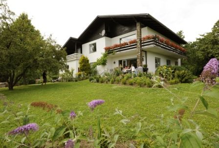 Dvoupatrový bílý dům s balkonem, obklopený zelenou zahradou s květinami a stromy.