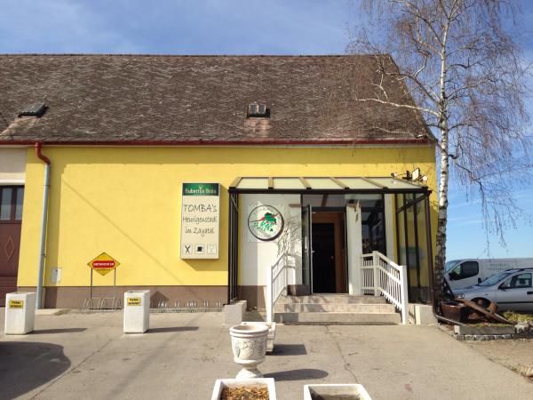 Žlutá budova s nápisem "Tomba's Heurigenschenke". Vchod se schody a prosklenou střechou.
