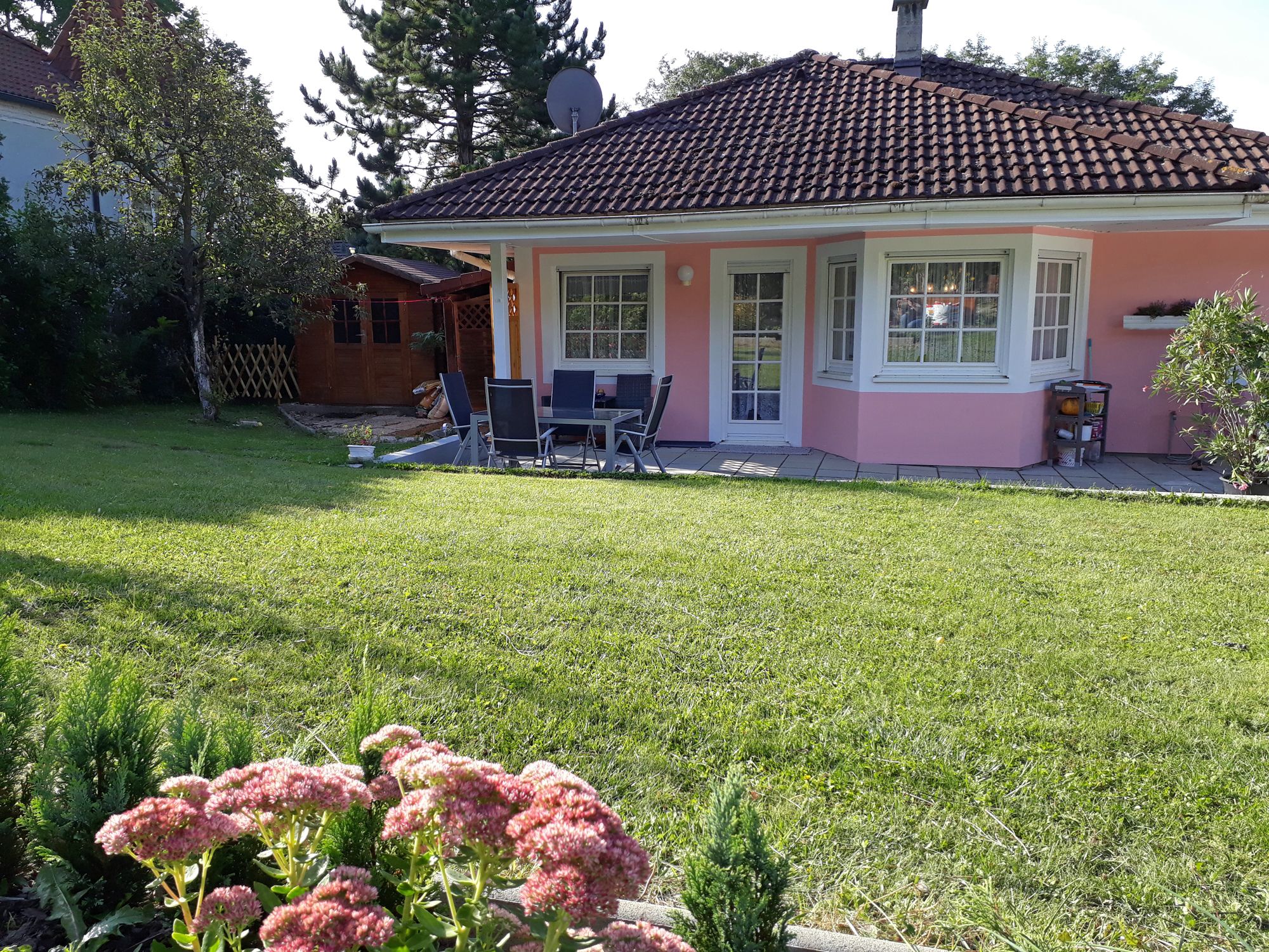 Růžový dům se zahradou, terasou a zahradním nábytkem, obklopený stromy a květinami.