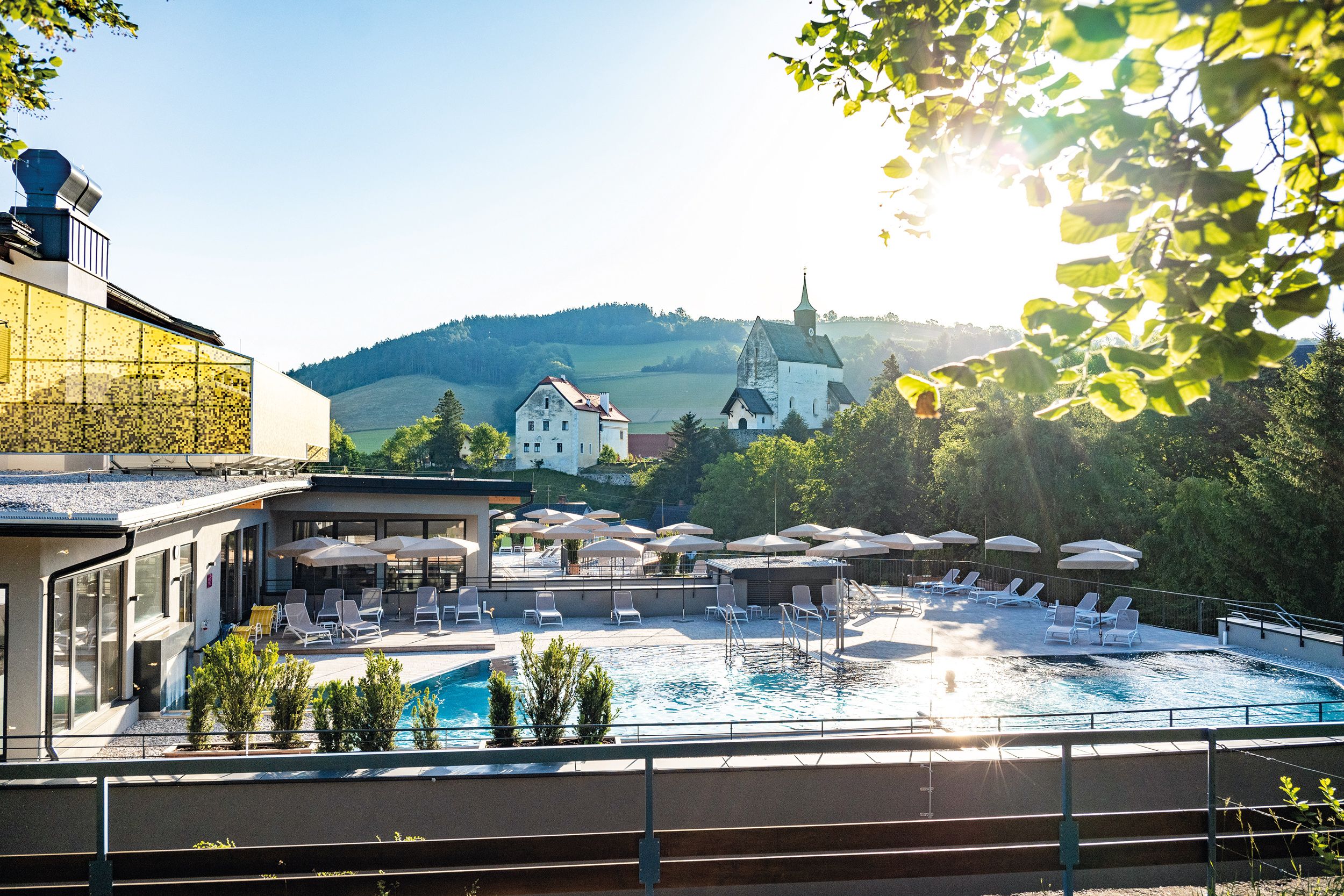 Venkovní areál hotelu s bazénem, slunečníky a lehátky, obklopený přírodou a kostelem v pozadí.