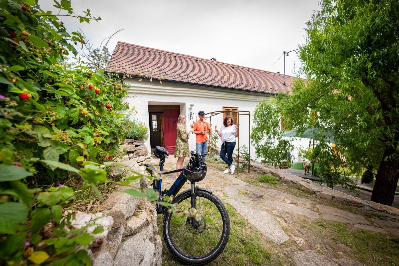 Tři lidé stojí před bílým domem s červenou střechou, v popředí je kolo.