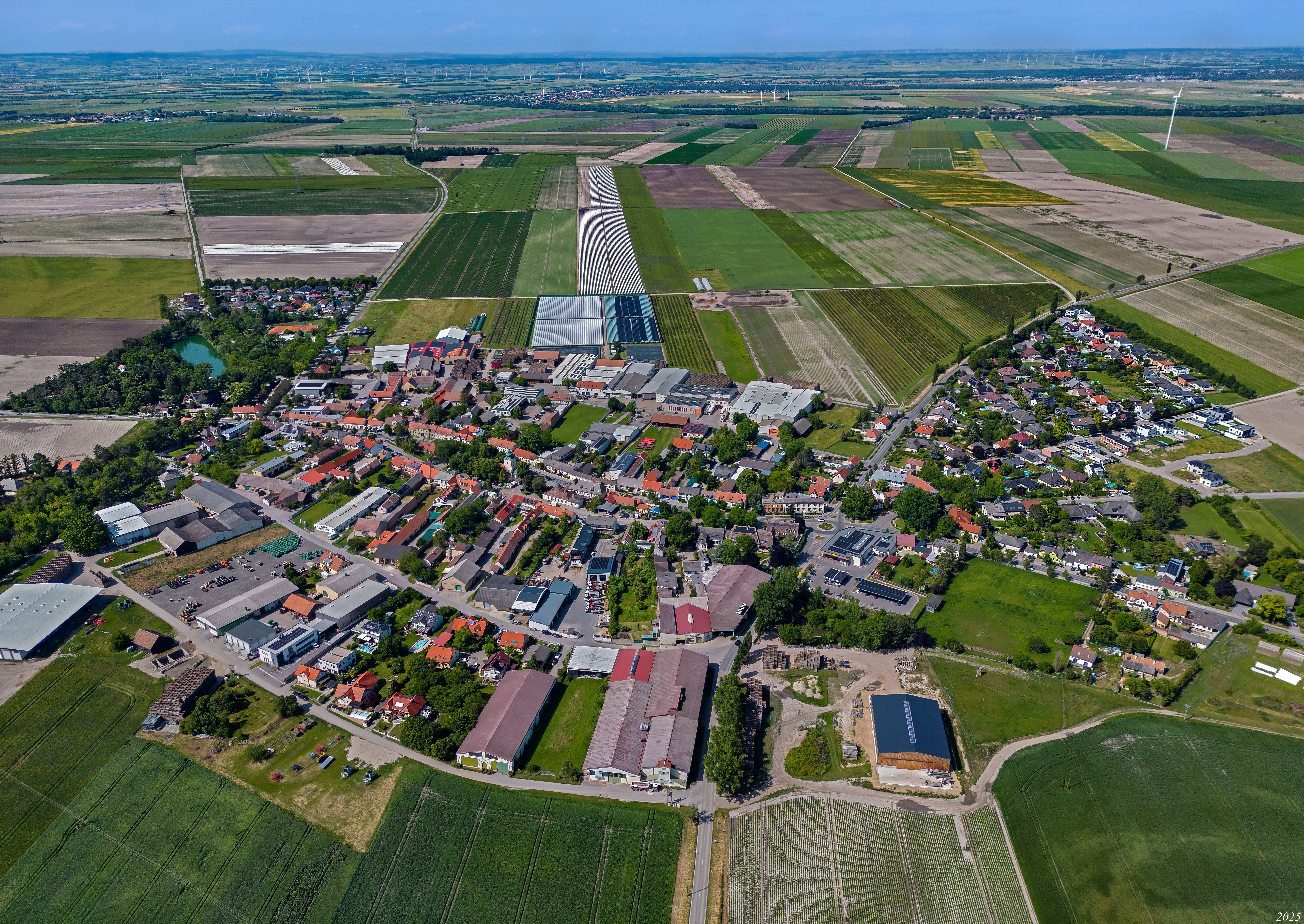 Letecký snímek Raasdorfu s centrem obce, poli, skleníky a větrnými elektrárnami v okolí.