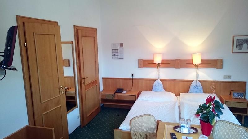 Hotelový pokoj se dvěma samostatnými postelemi, dřevěným nábytkem a zeleným kobercem.