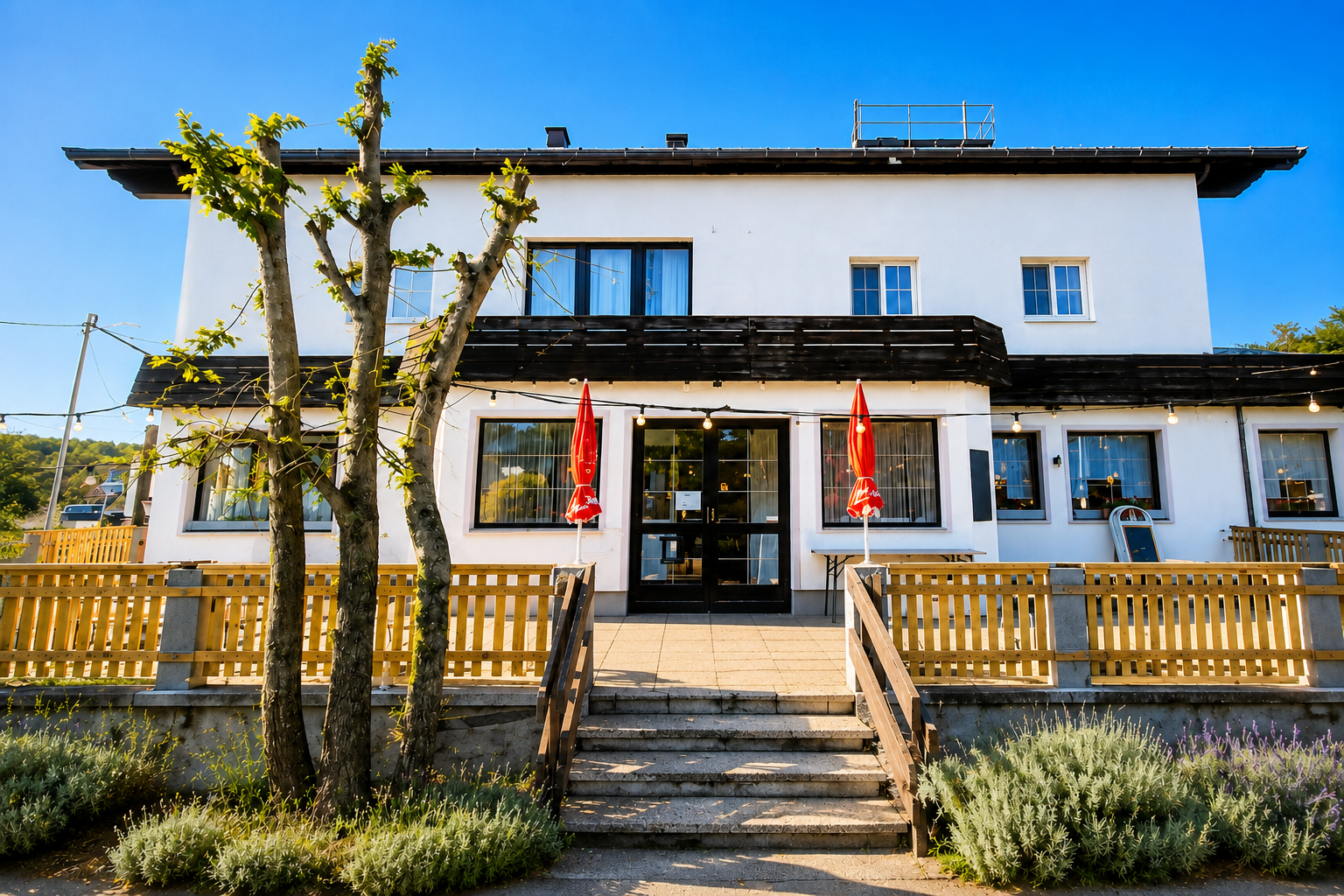 Vnější pohled na dvoupodlažní restauraci s bílými stěnami a červenými slunečníky před vchodem.