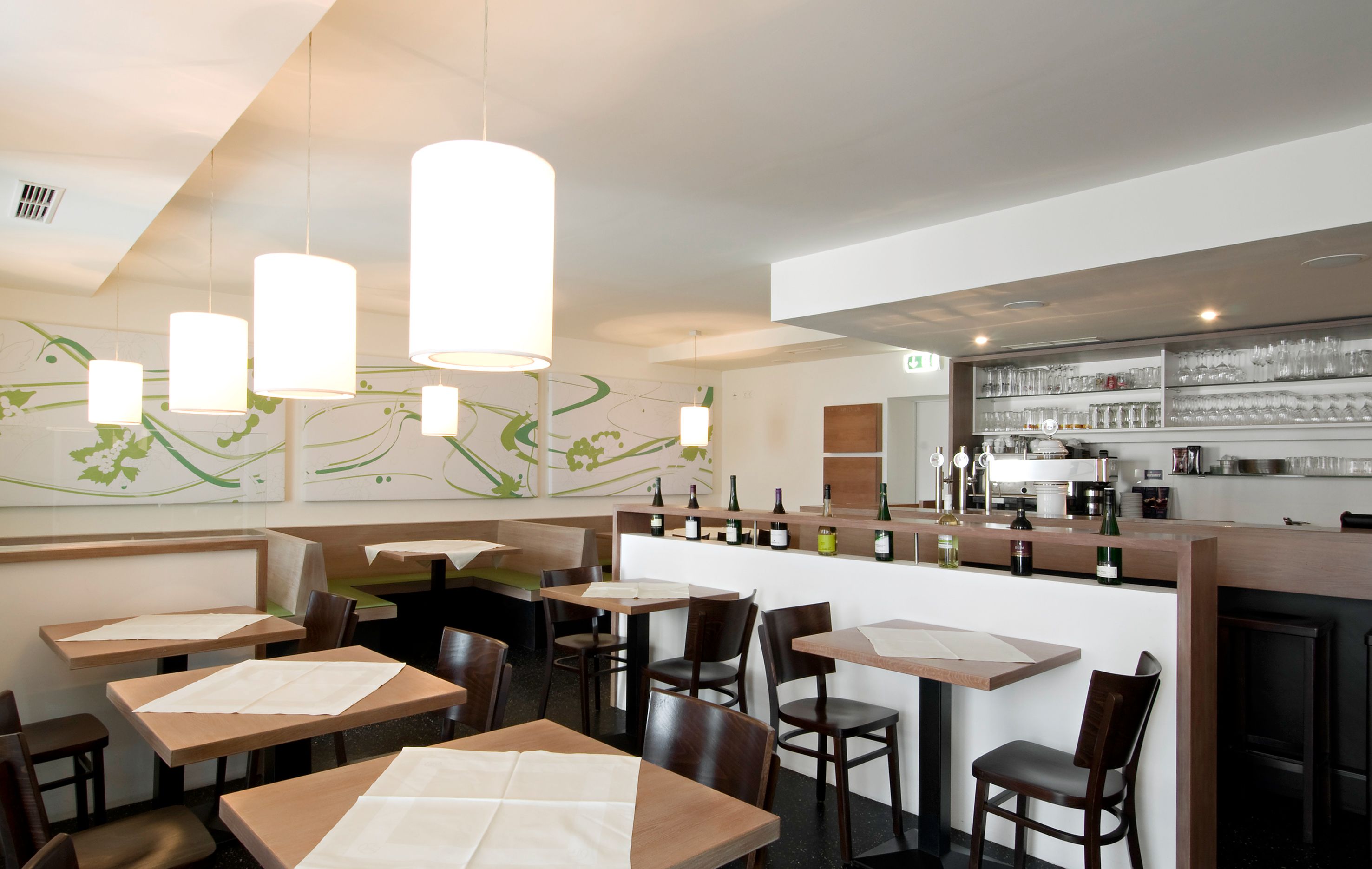 Pohled do interiéru moderní restaurace s dřevěnými stoly, židlemi a barem s lahvemi a sklenicemi.