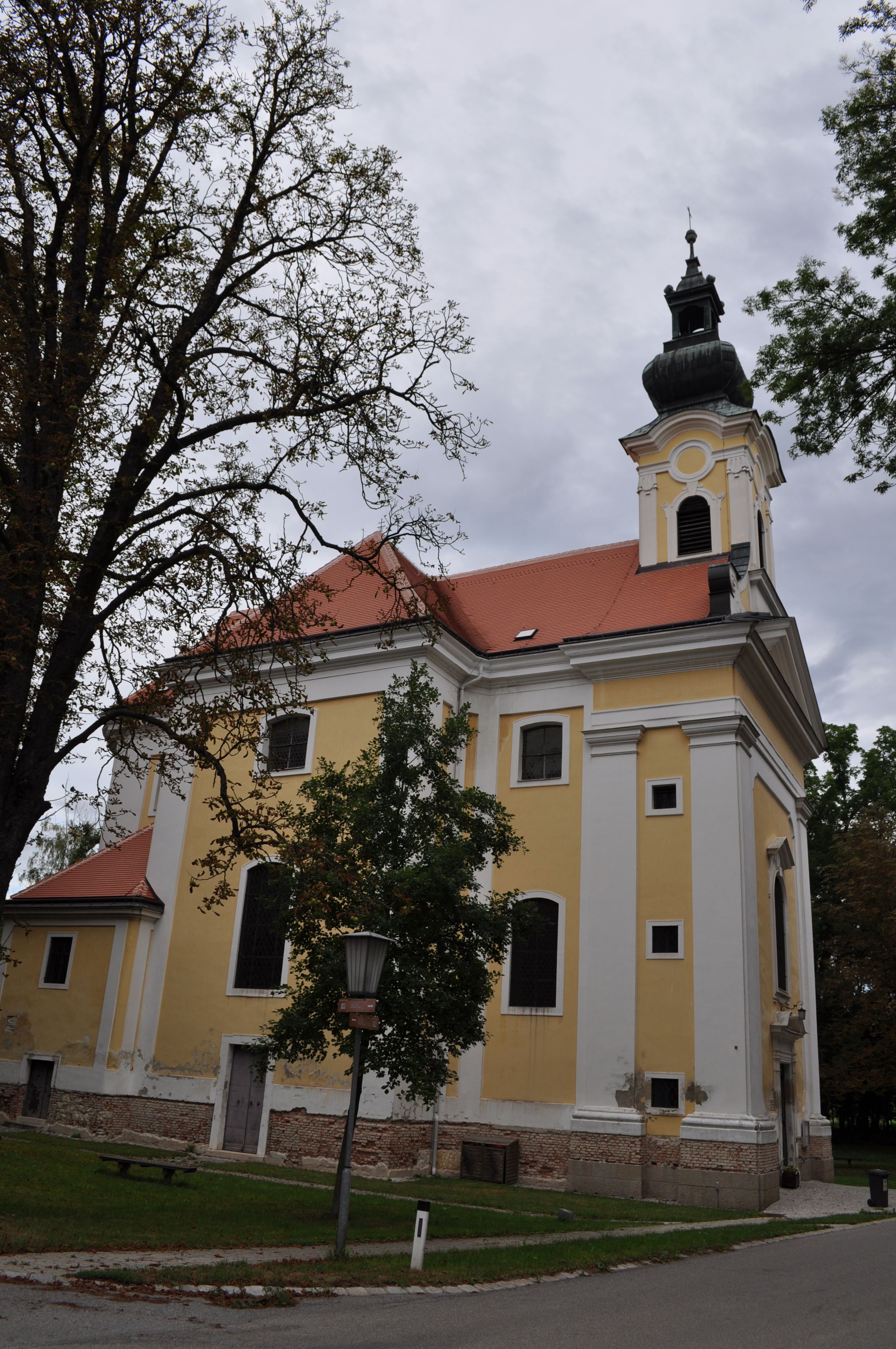 Žlutý kostel s červenou střechou a věží, obklopený stromy.